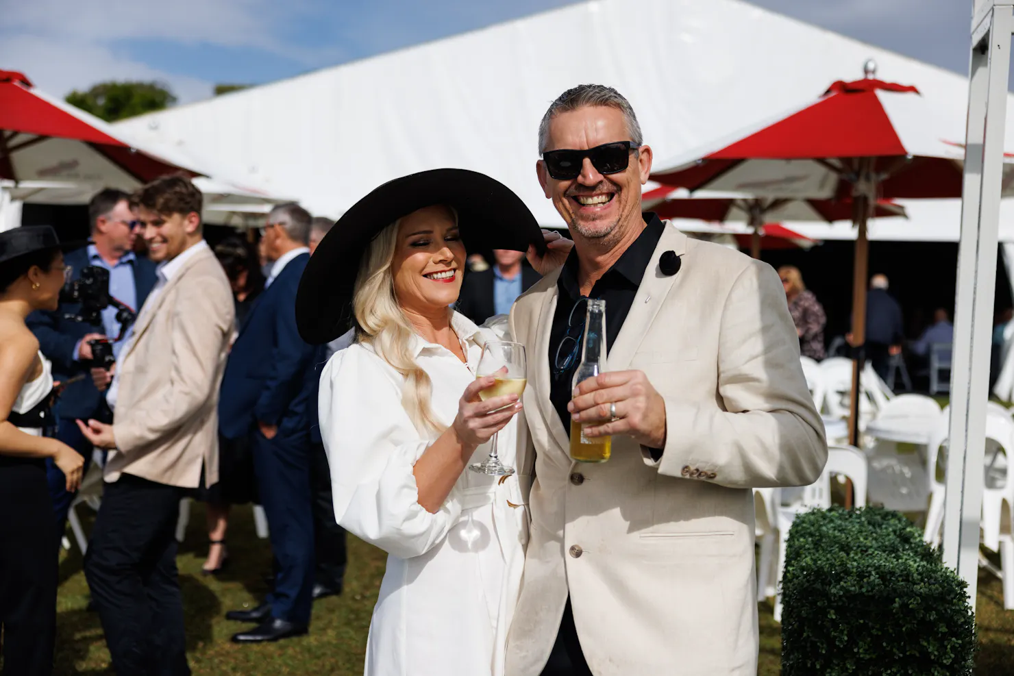 male and female smiling at the camera holding up drinks in front of a marquee