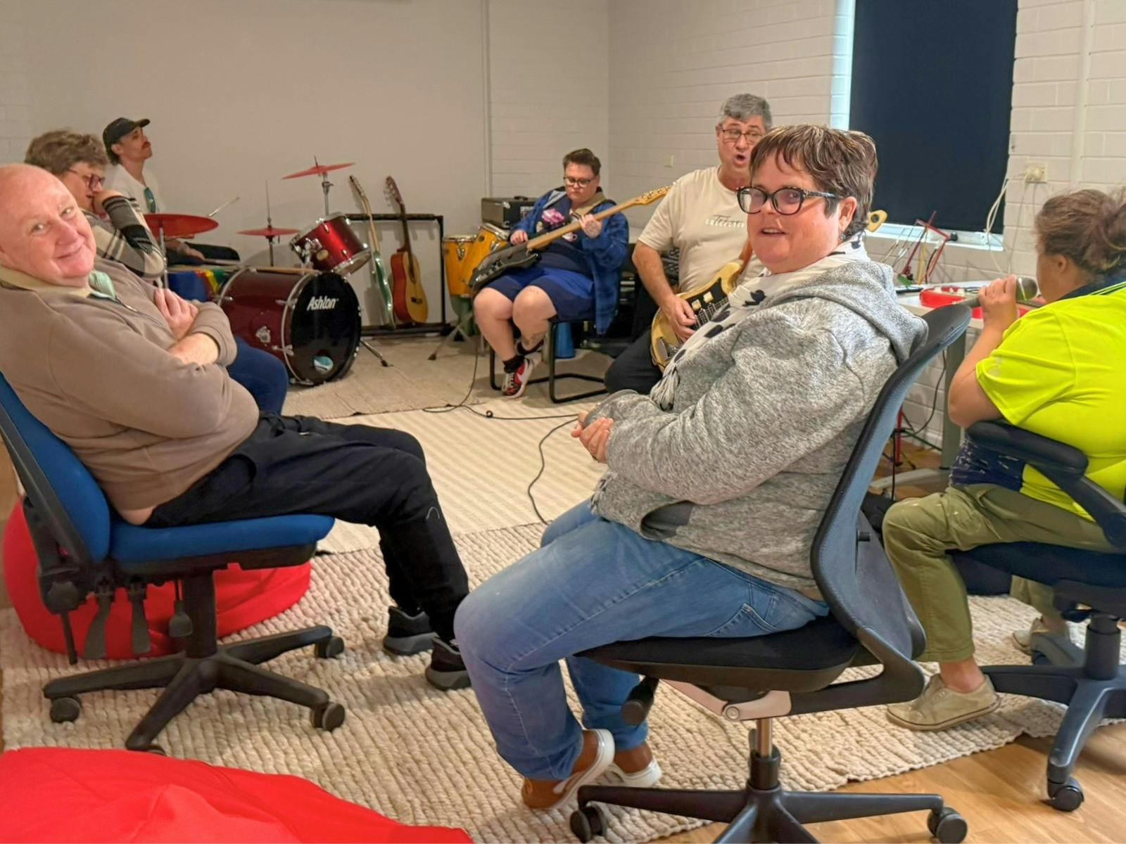 Participants sit in a circle while playing instruments including electric guitar, bass guitar and dr