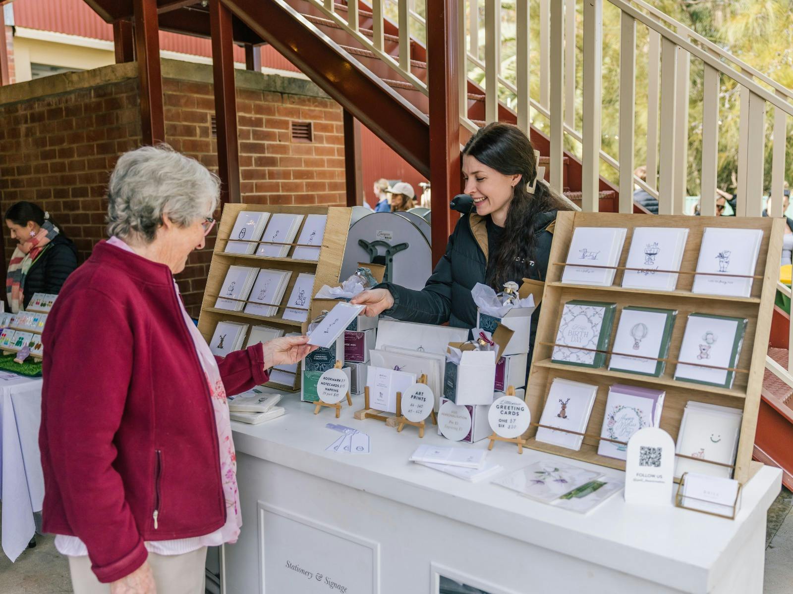 A stationary market stall, the artist is smiling and handing over a handmade card to the customer.