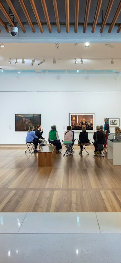 A group of people are seated in a brightly lit gallery, discussing portraiture with a guide.
