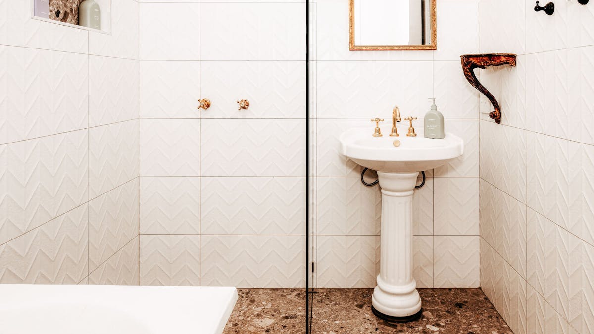 A white tiled walk in shower and white pedastal basin with gold rimmed mirror and gold tapware