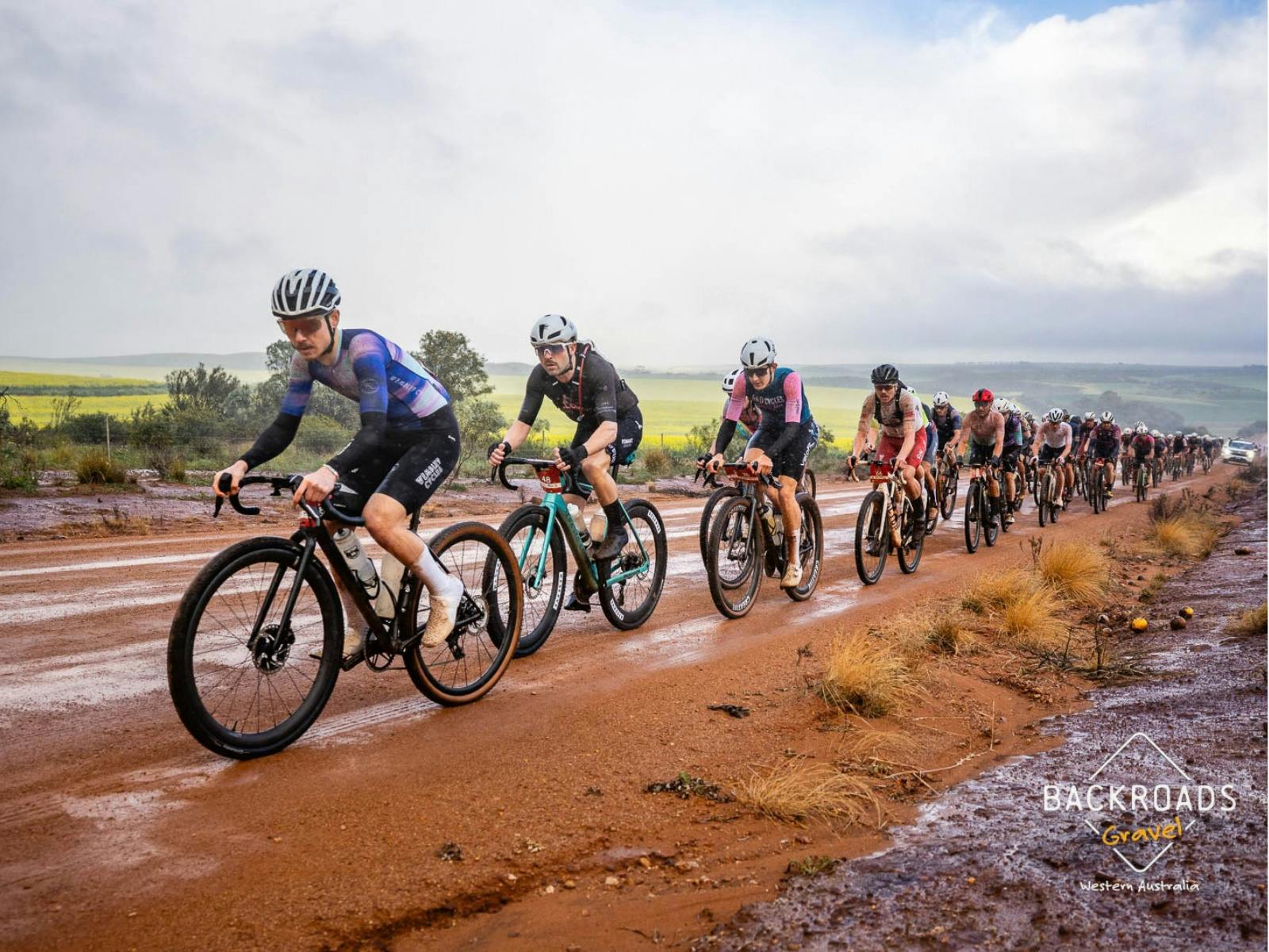 Group of gravel cyclists riding along a wet red dirt road in Chapman Valley.