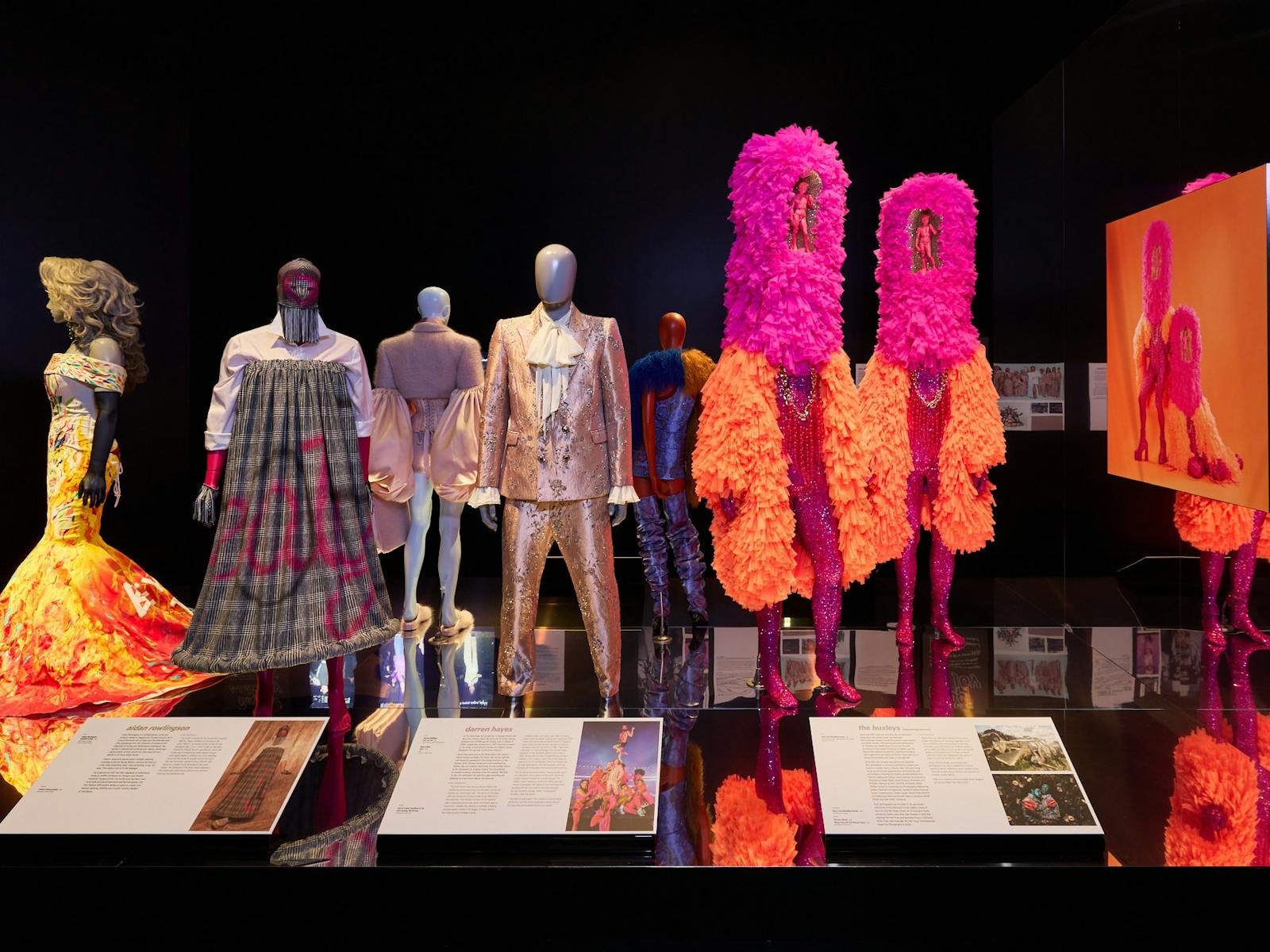 Vibrantly dressed mannequins are positioned along a reflective black platform. Vibrantly dressed mannequins are positioned along a reflective black platform.