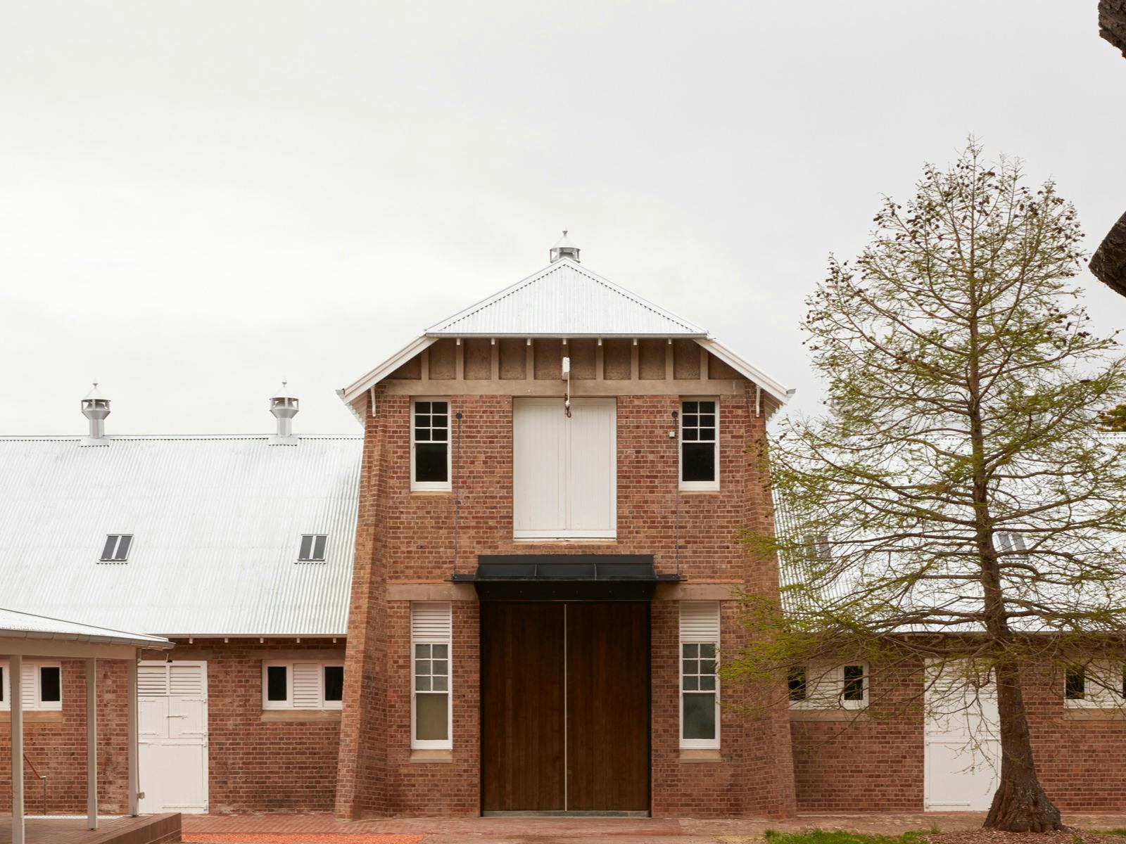 A large old converted dairy stands in front of a grey sky next to a tall tree