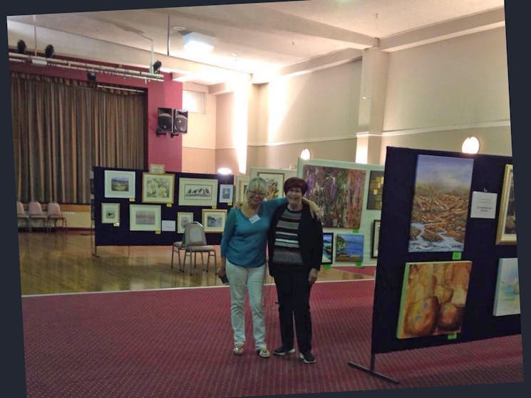 two ladies standing in the hall with art work on display around them.