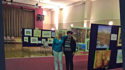 two ladies standing in the hall with art work on display around them.
