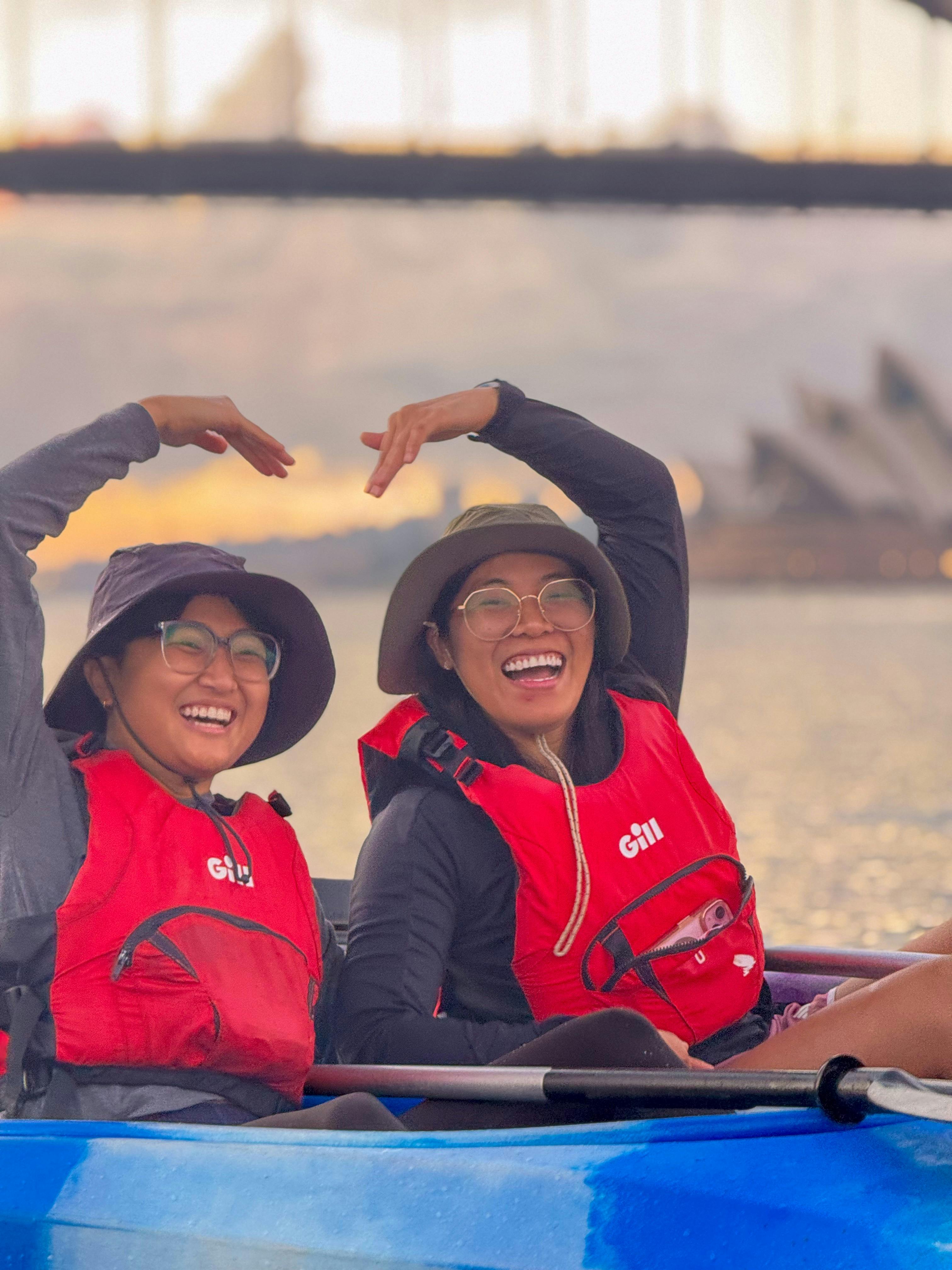 Two ladies use their hands to impersonate a bridge over them from their kayaks with Opera House.