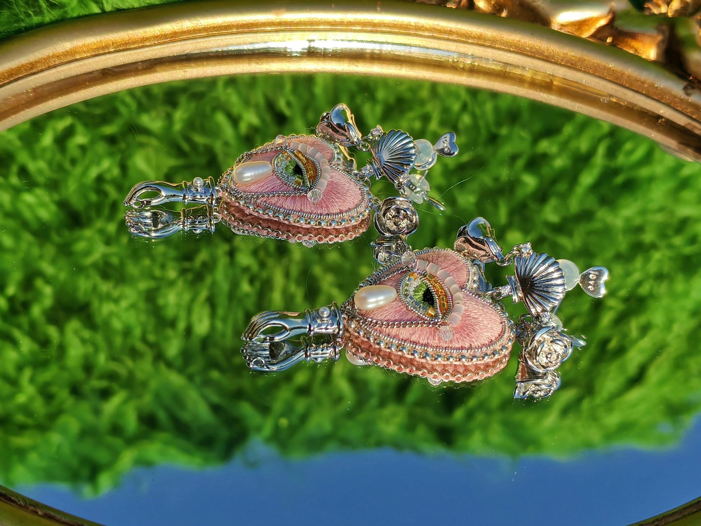 Two pink embroidered eye-shaped jewellery pieces reflected in a round mirror.
