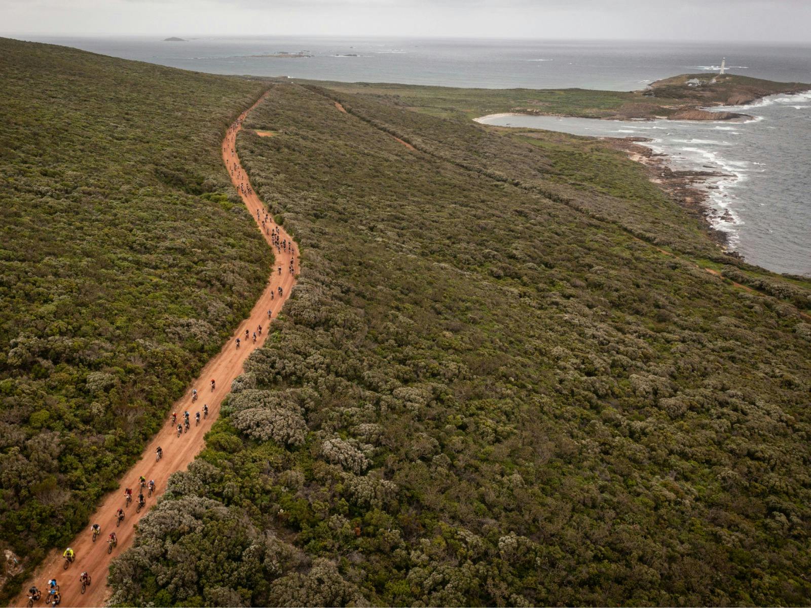 Drone aerial shot of mountain bikers on red gravel road amongst bushland, with coast in background