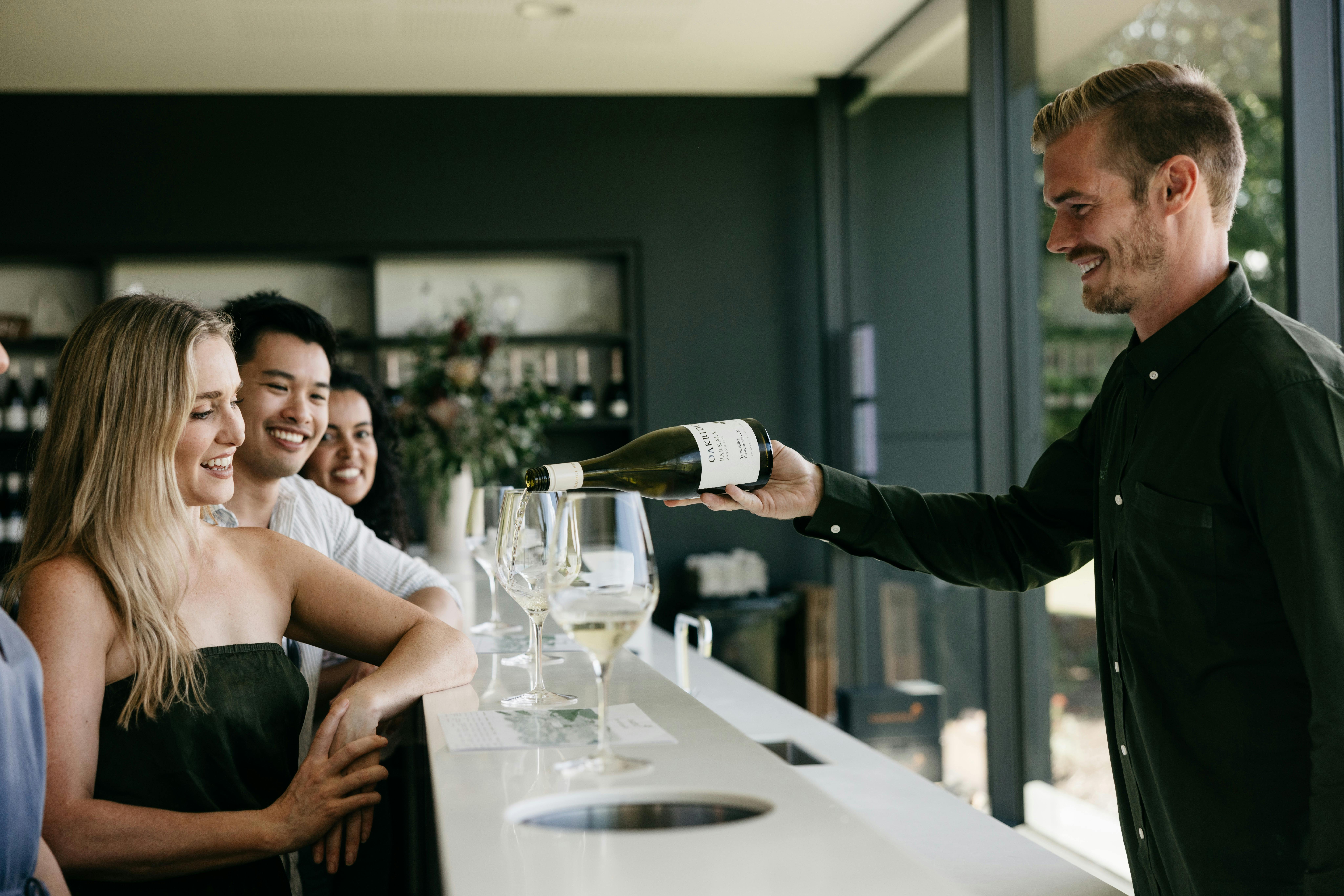 3 guests sit at the tasting bar while a smiling staff member pours Oakridge Chardonnay