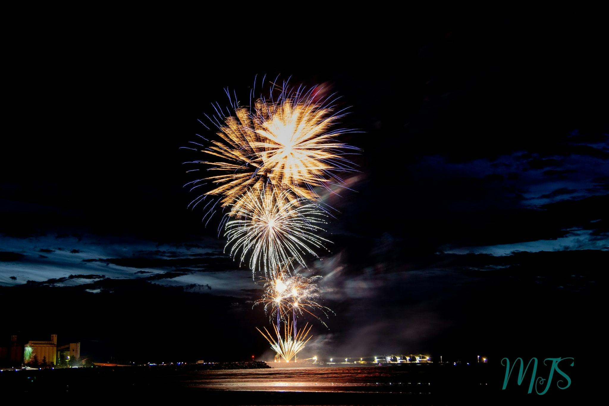 4 large golden fireworks with speckles of purple and blue exploding over the night sky and sea.