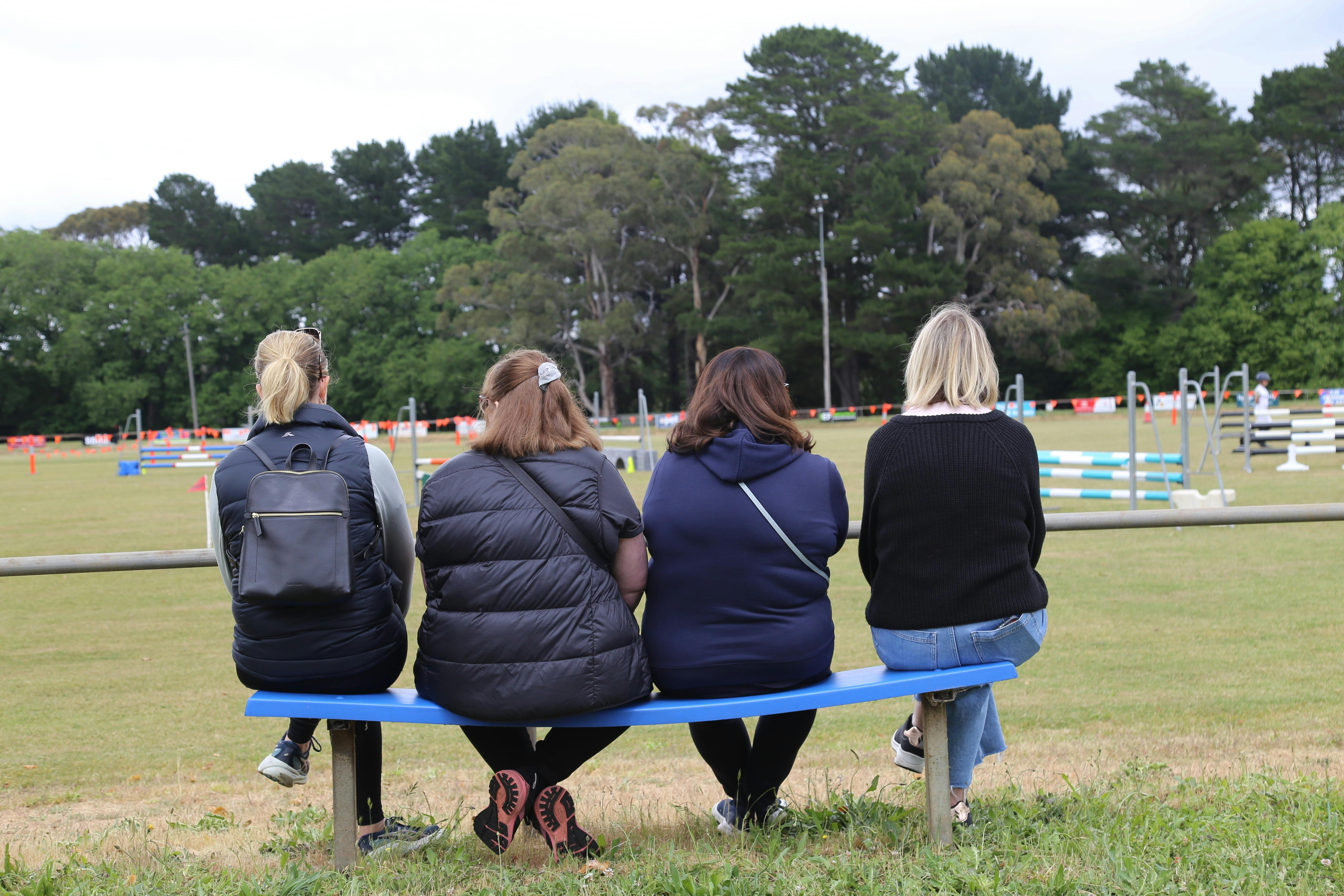 Backs of 4 women sitting on blue bench watching Show Jumping event