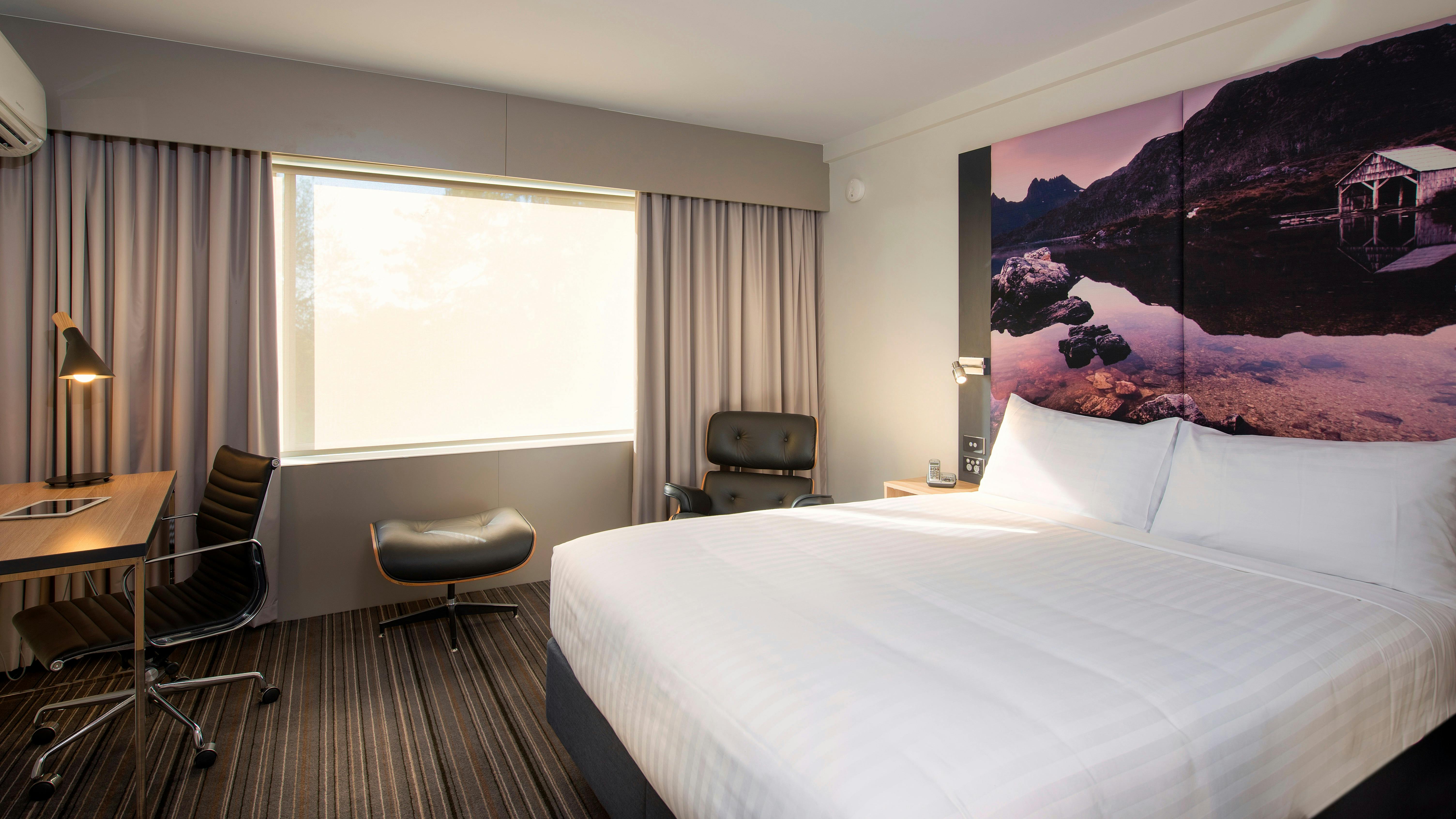 Guest accommodation room, with a queen bed, chair and footstool, desk and chair, window with blind.