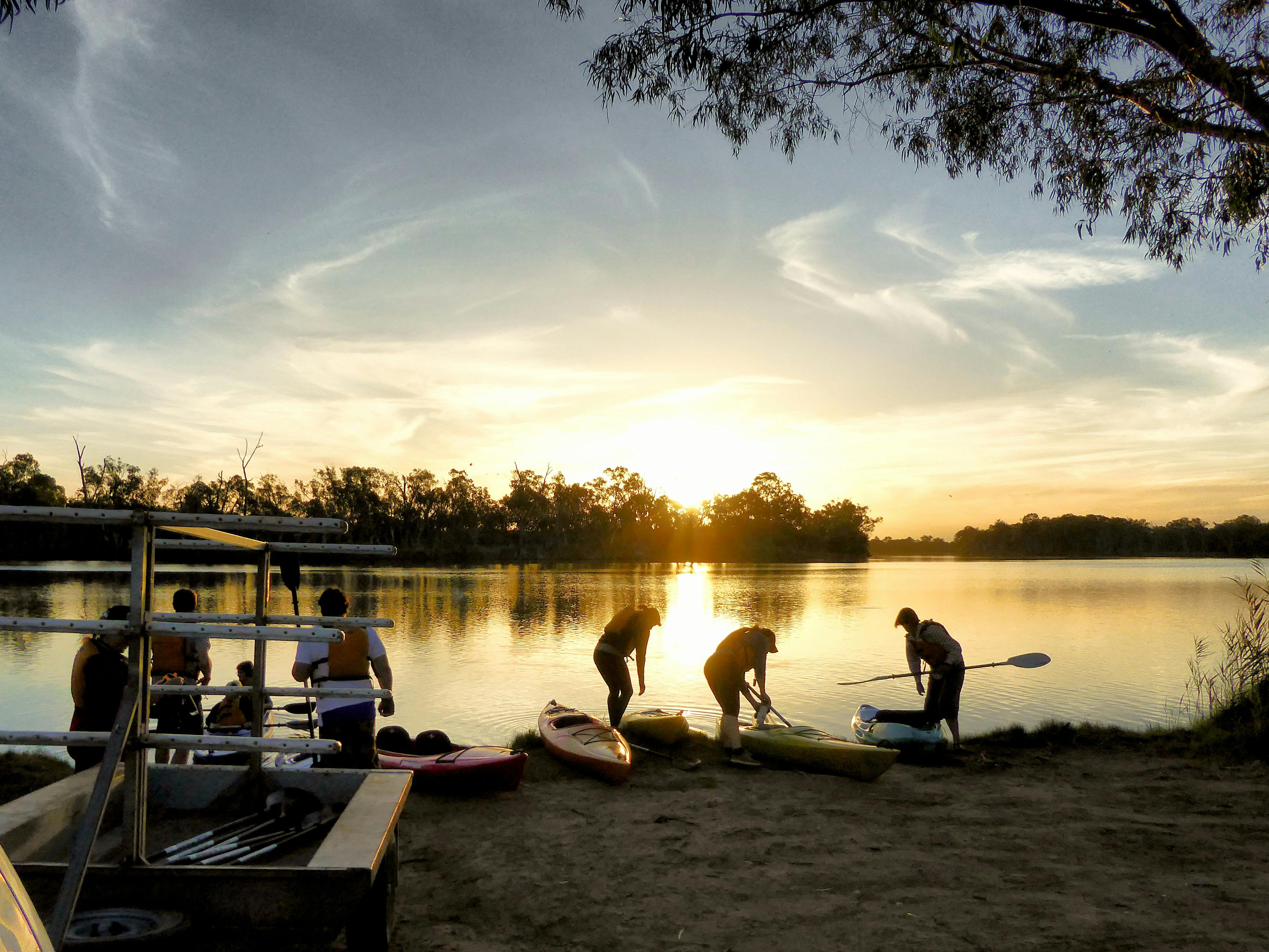 3 kayakers step into their kayaks on the waters edge as the sun sets behind them.  kayak trailer nea
