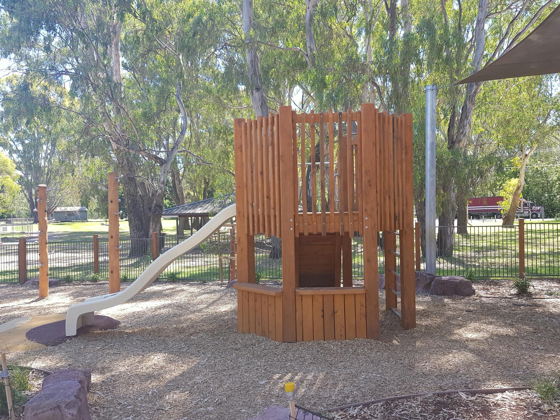 Childrens playground, wooden structure with steps to slide, leafy gum trees, shade sails