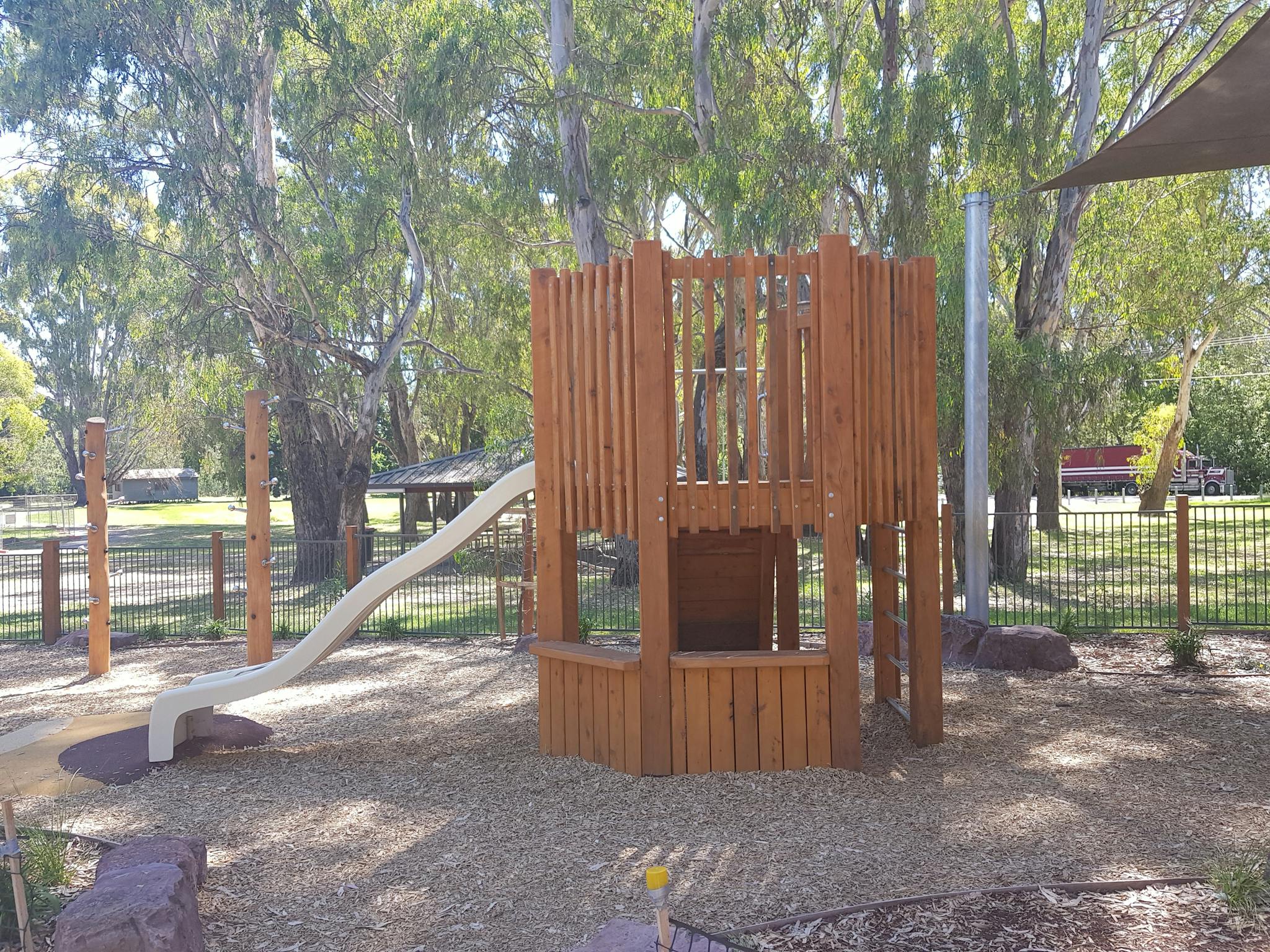 Childrens playground, wooden structure with steps to slide, leafy gum trees, shade sails