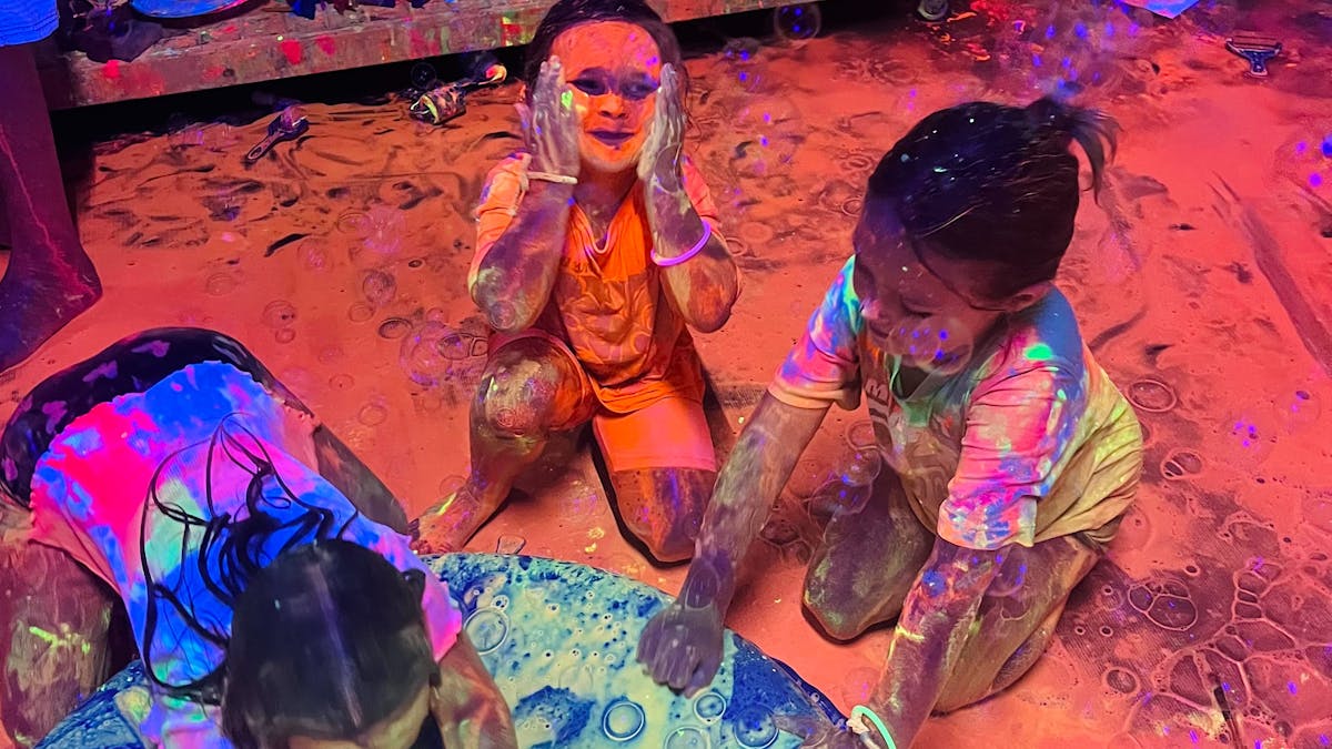 A little girl covering her face in glowing paint at Messy Glow Co UV Paint Room