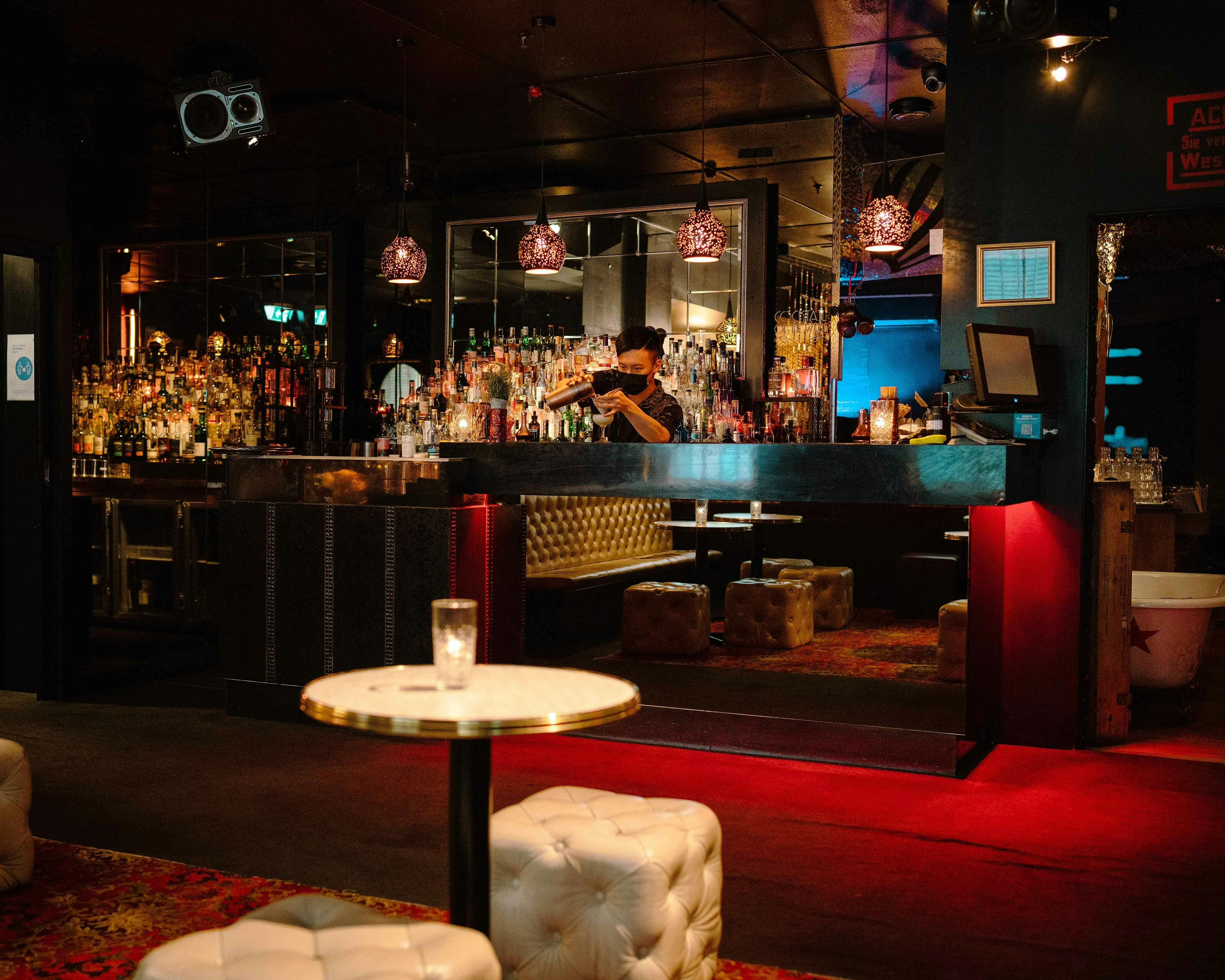 Inside a cocktail bar.  There is red carpet and a cream table and stool.  A bar is at the back.