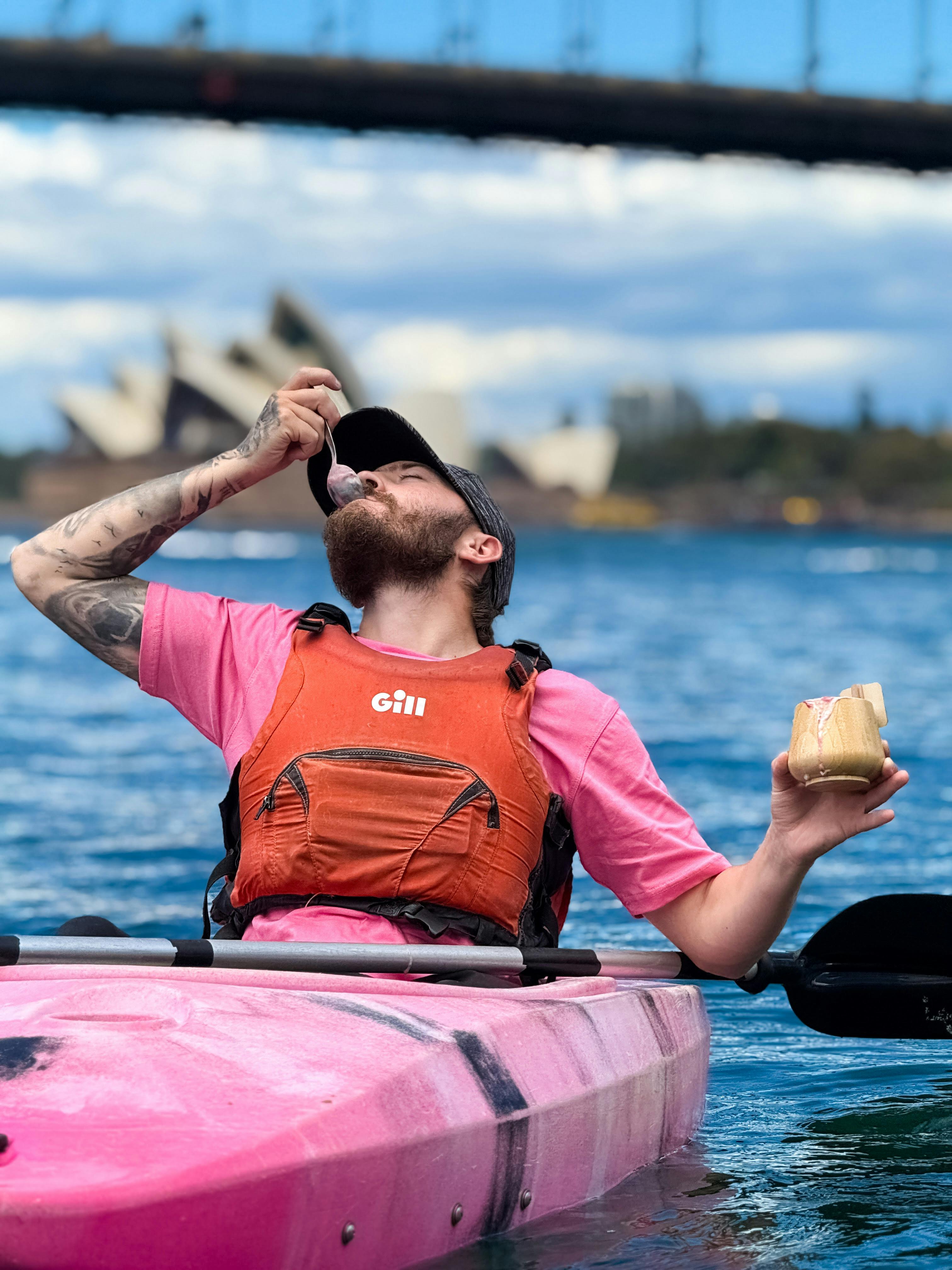 Man in pink shirt eating gelato ice cream from kayak with Sydney Opera House in background.