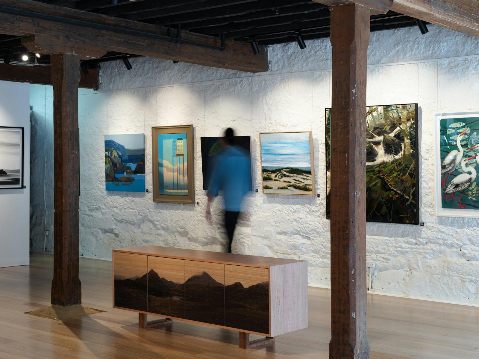 Image of inside Handmark, showing person walking past exhition works and viewing paintings.