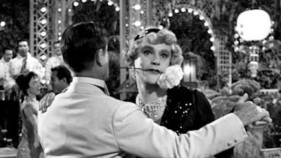 Black and white still of two men dancing, one dressed as a woman with a flower in his mouth