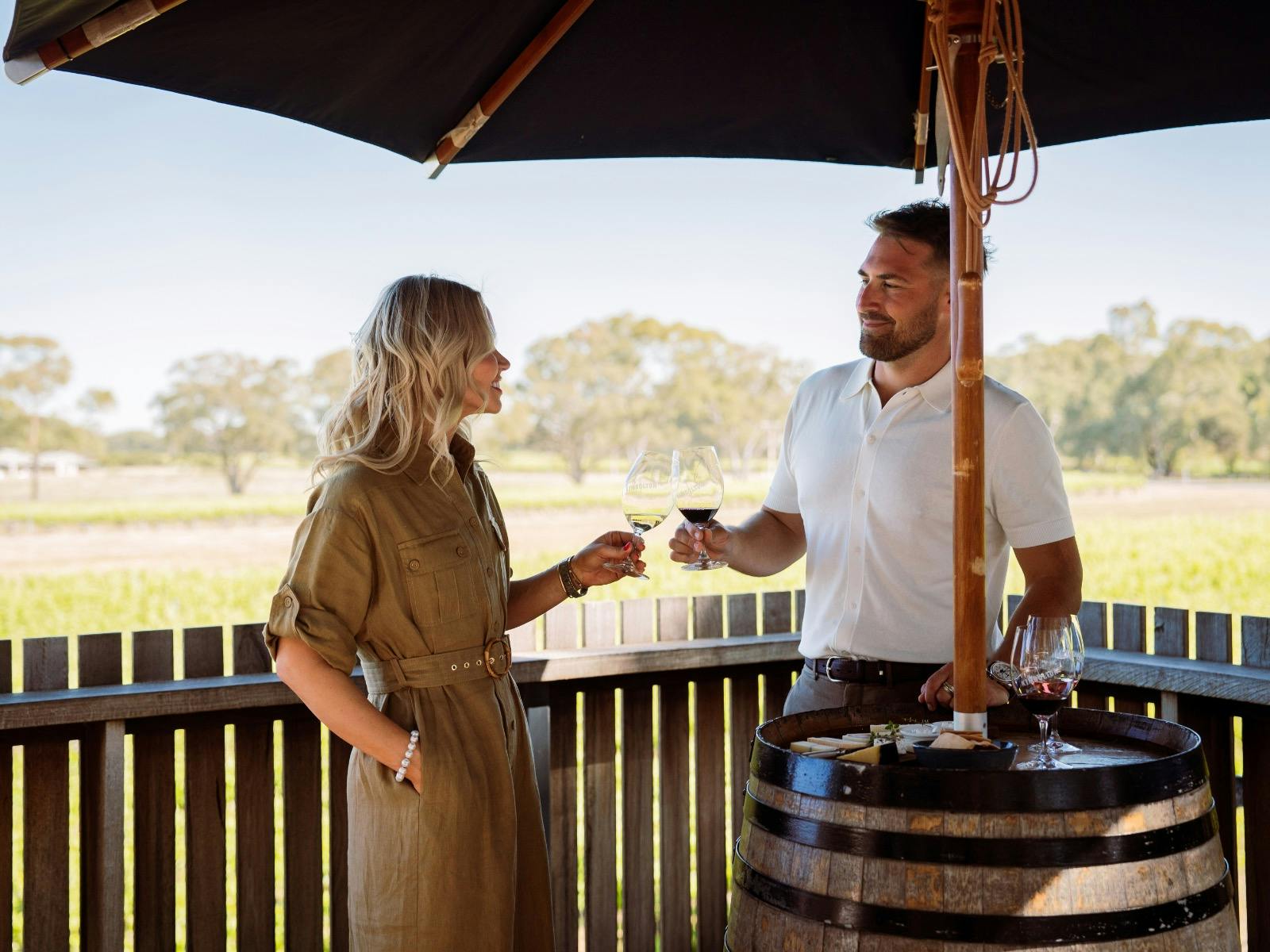 A couple stand on a rooftop clinking glasses. There is a cheese board on a barrel and vine views.