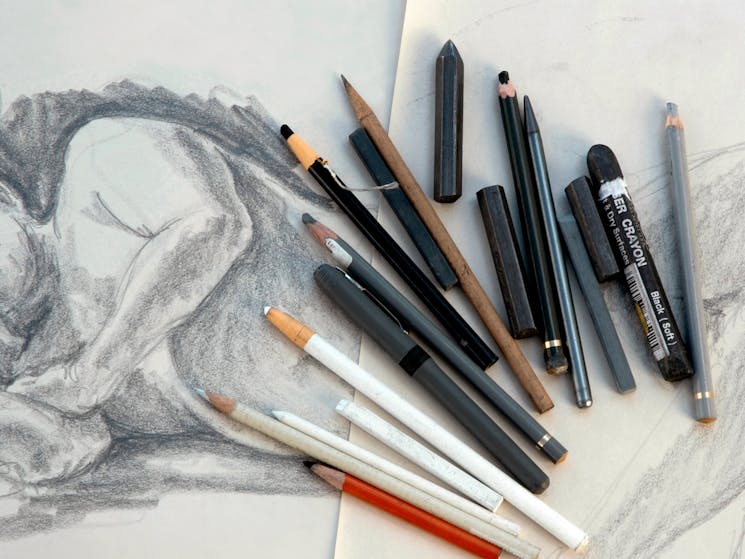 Charcoal, pencils, in different monochromatic shades splayed out over a sketch of a reclining figure