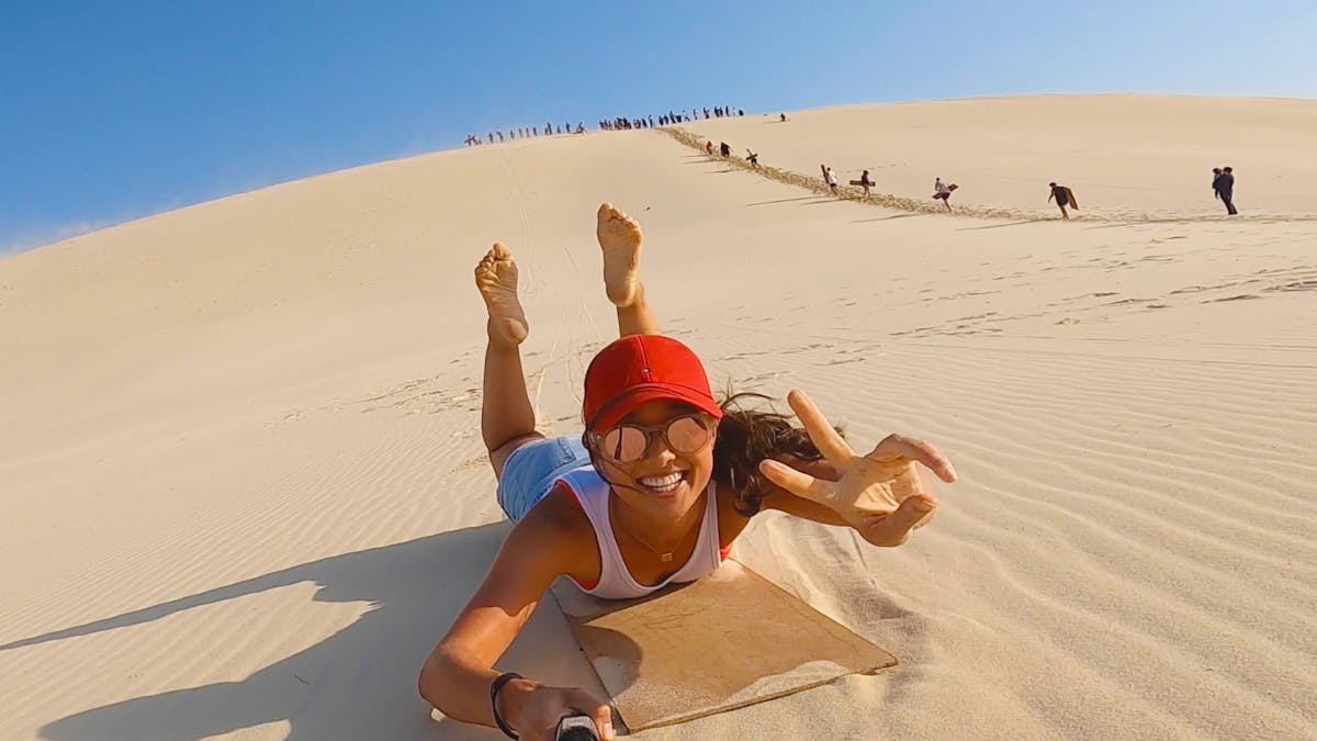 Women on a sandboard riding down a sand due, holding a gopro in one hand
