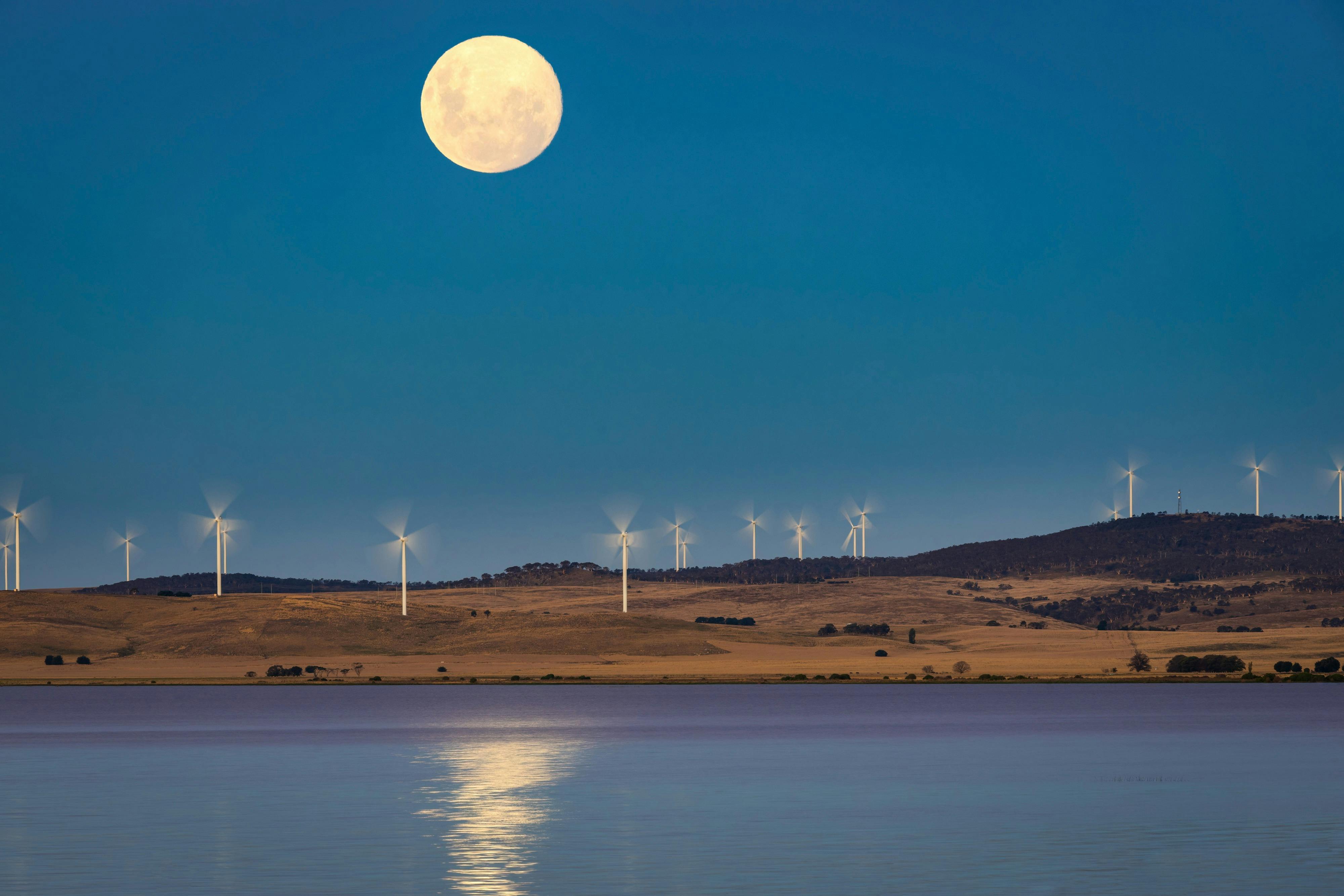 A bright full moon hangs above a dark open landscape, with wind turbines along the horizon and water