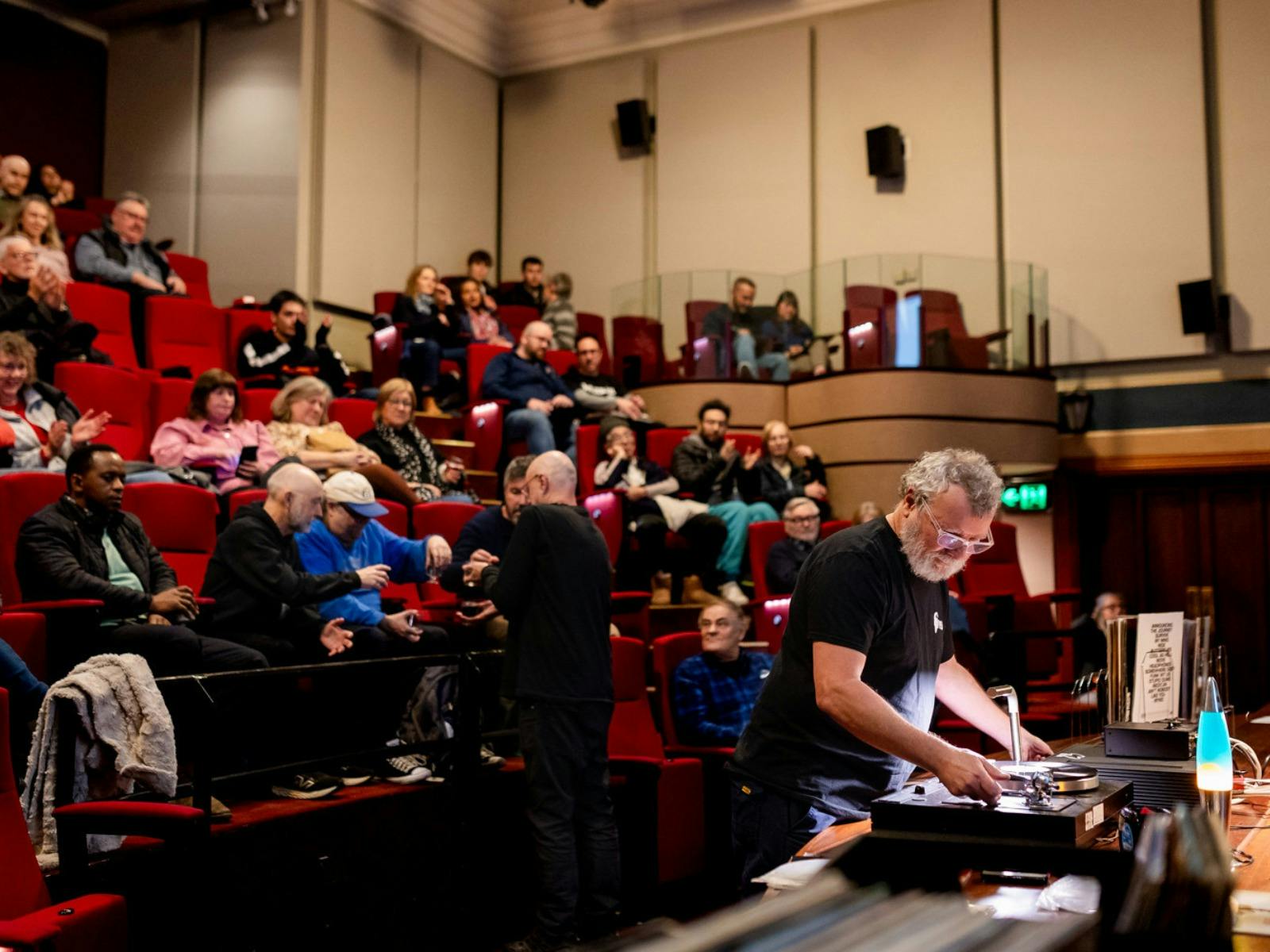 Audience in a theatre and a man at the front preparing to play a vinyl