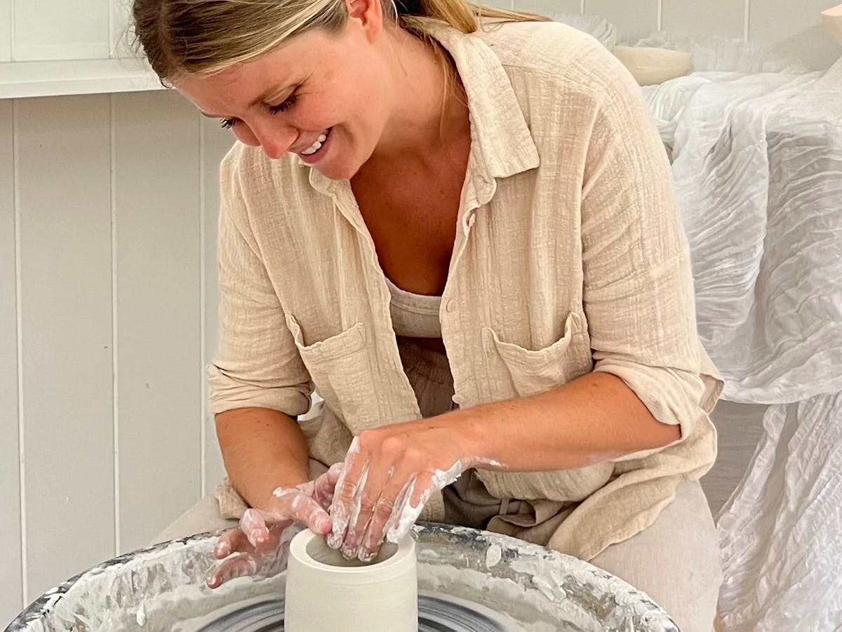 Smiling woman creating pottery on a wheel in a light-filled ceramic studio