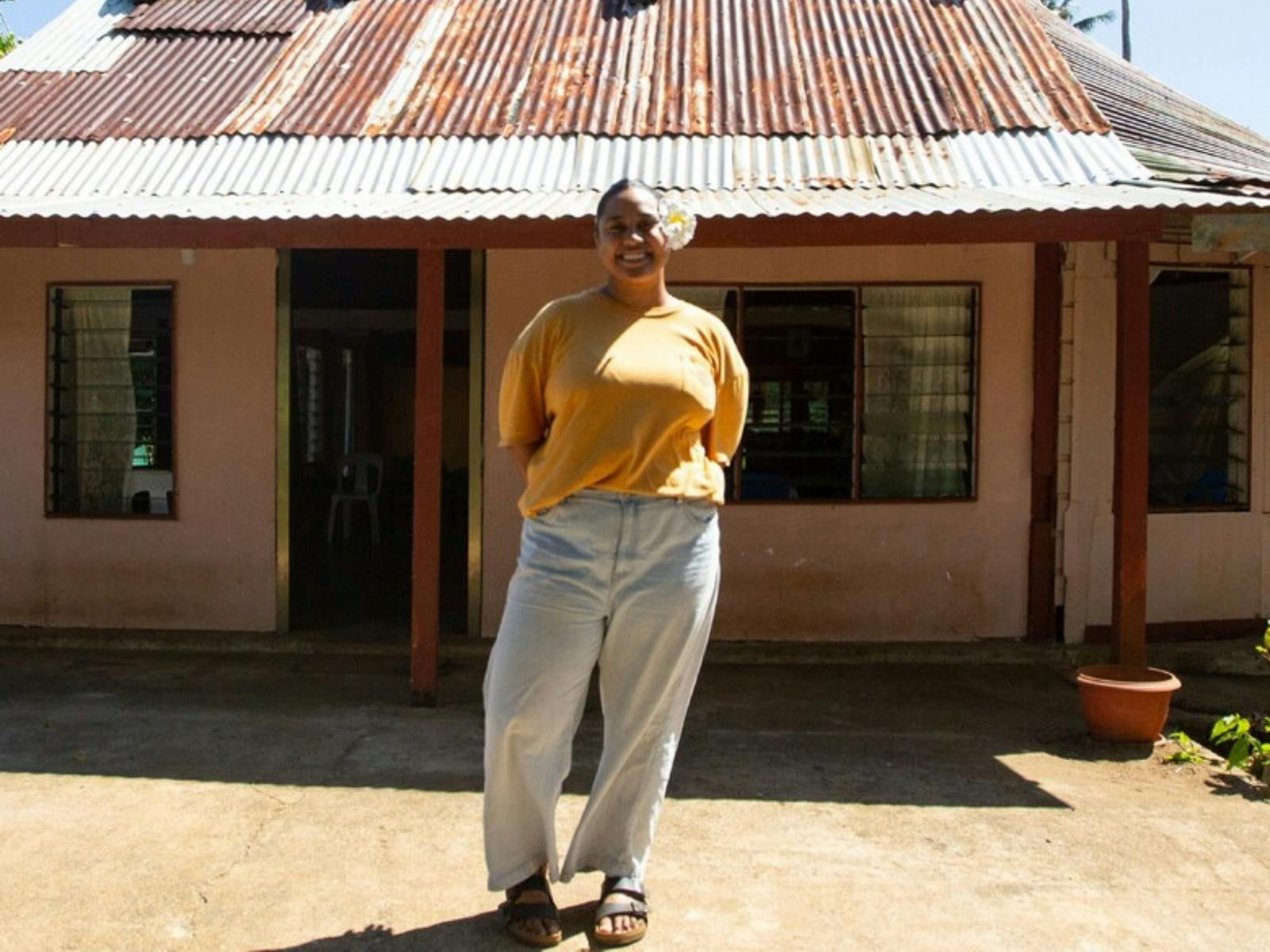 A smiling woman in a yellow shirt and light blue pants stands in front of a house.