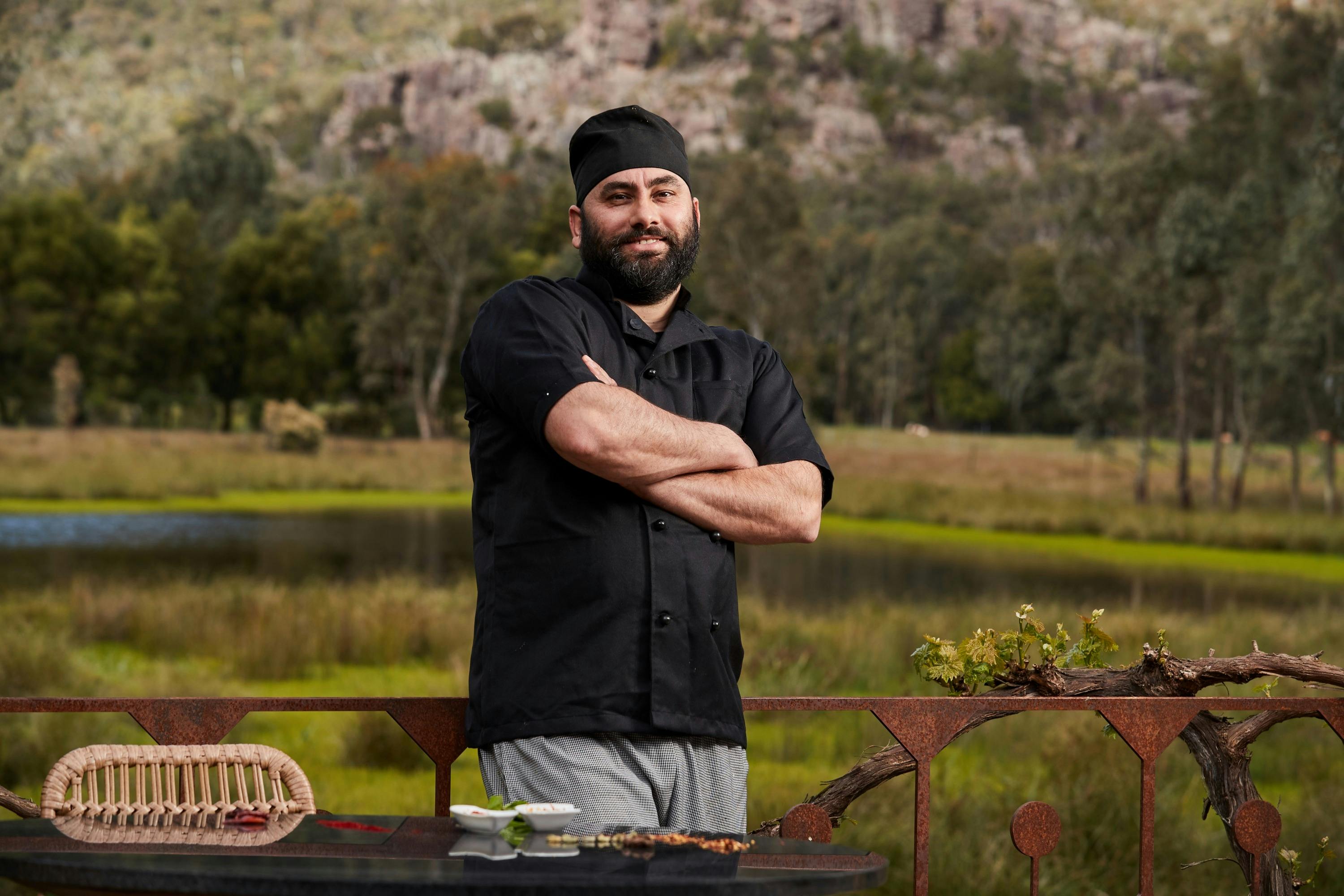 A chef standing next to a table outside with a pond and mountains in the background.