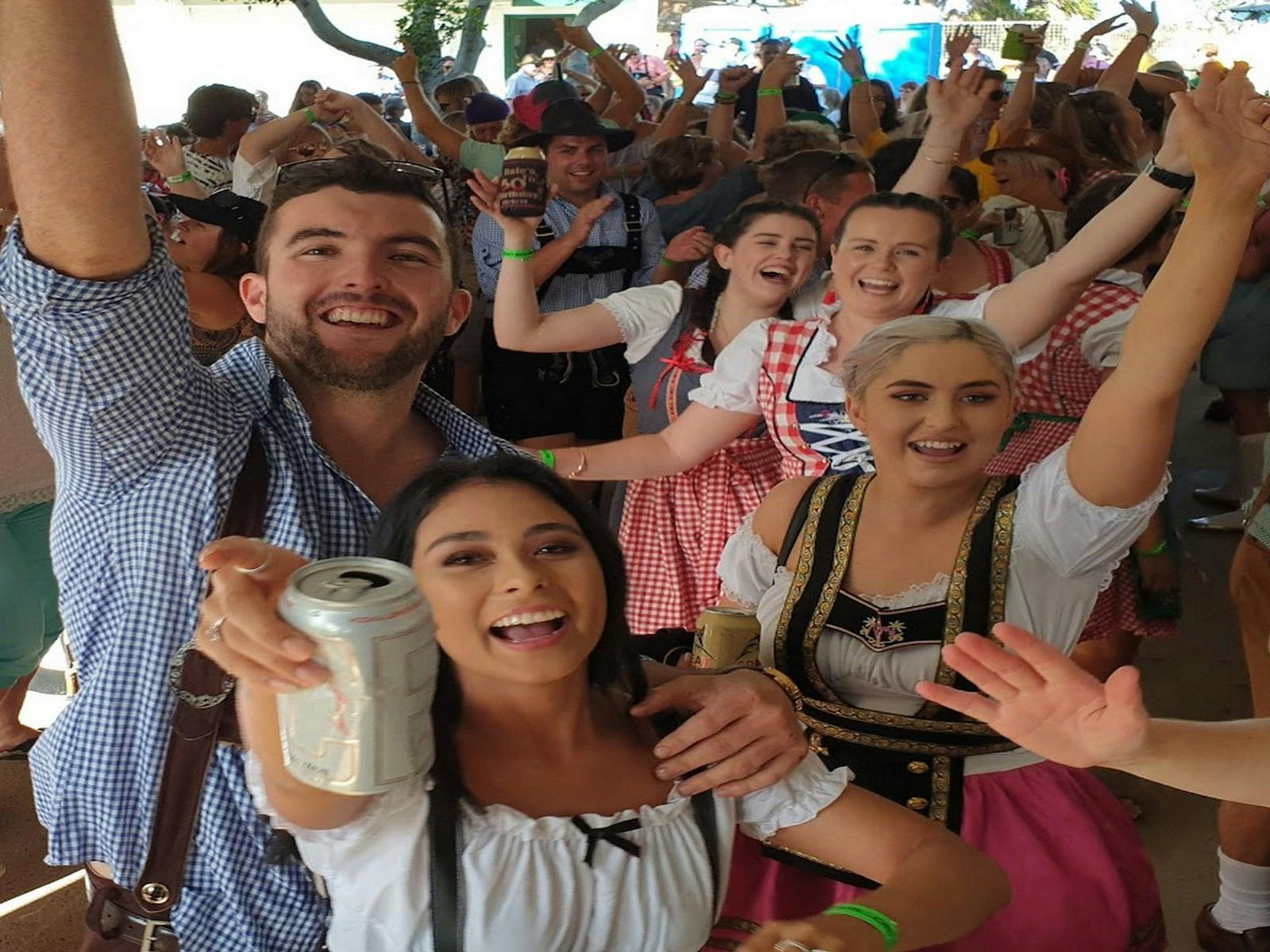 A young group of males and females in traditional Bavarian costume with raised arms holding drinks.