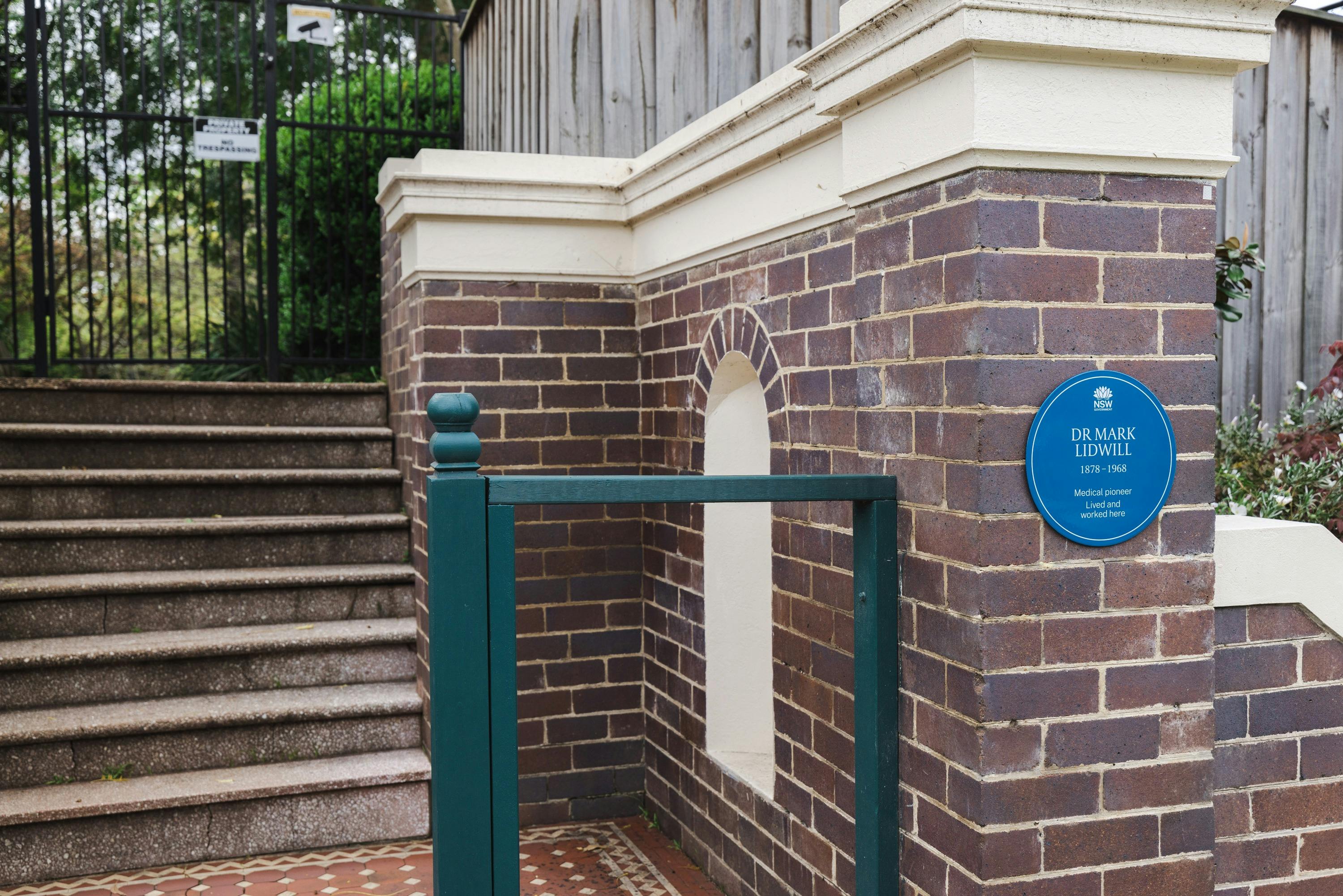 Blue plaque on a red/brown brick pillar, a federation green handrail next to steps leading to a gate