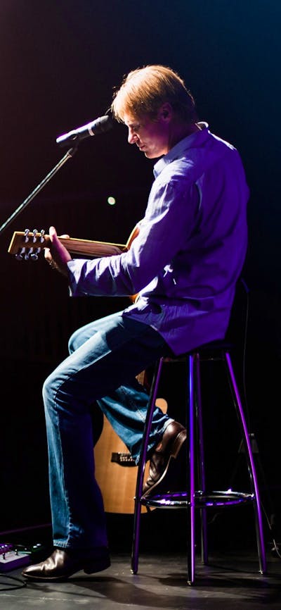 A man in jeans and a shirt sit on a bar stool in front of a microphone on stage, holding a guitar