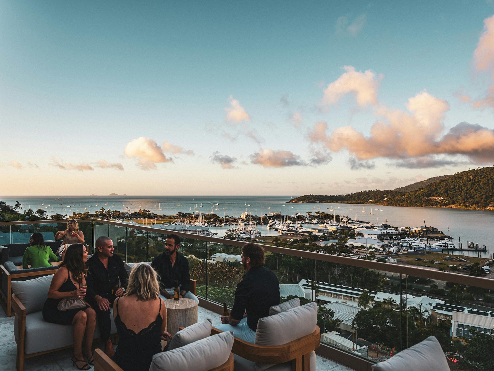 People socializing in lounge chairs on Elementa Whitsundays rooftop, overlooking the port and ocean. People socializing in lounge chairs on Elementa Whitsundays rooftop, overlooking the port and ocean.
