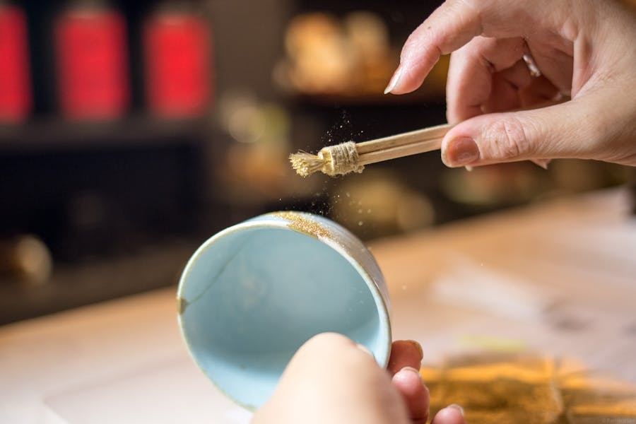 Picture shows one hand holding a blue bowl and the other sprinkling gold dust from a brush.