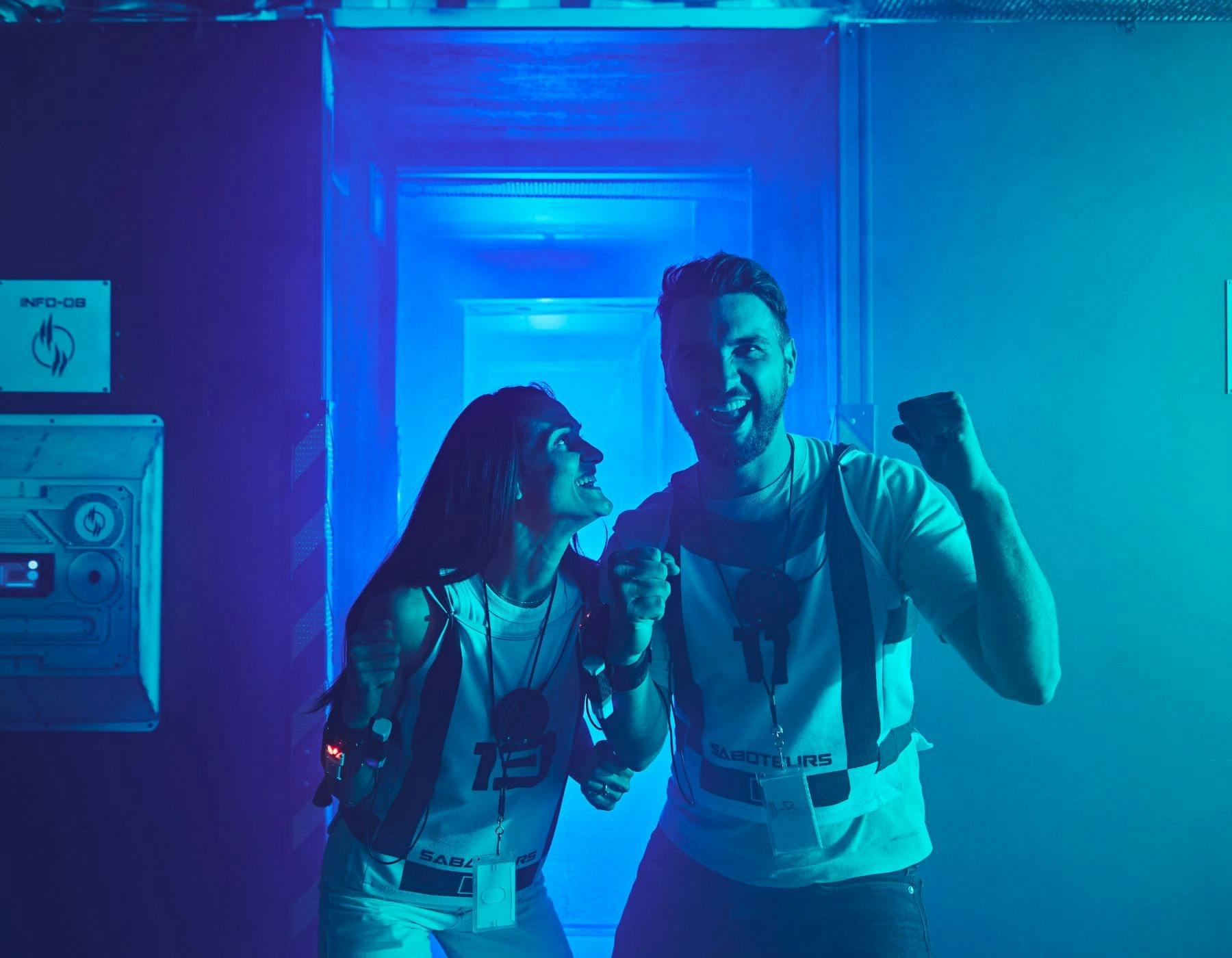 A man and woman in Saboteurs vests celebrate and laugh together in a blue-lit corridor.
