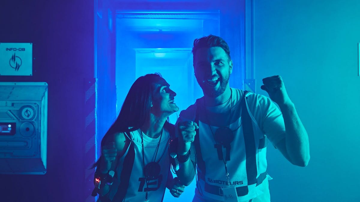 A man and woman in Saboteurs vests celebrate and laugh together in a blue-lit corridor.