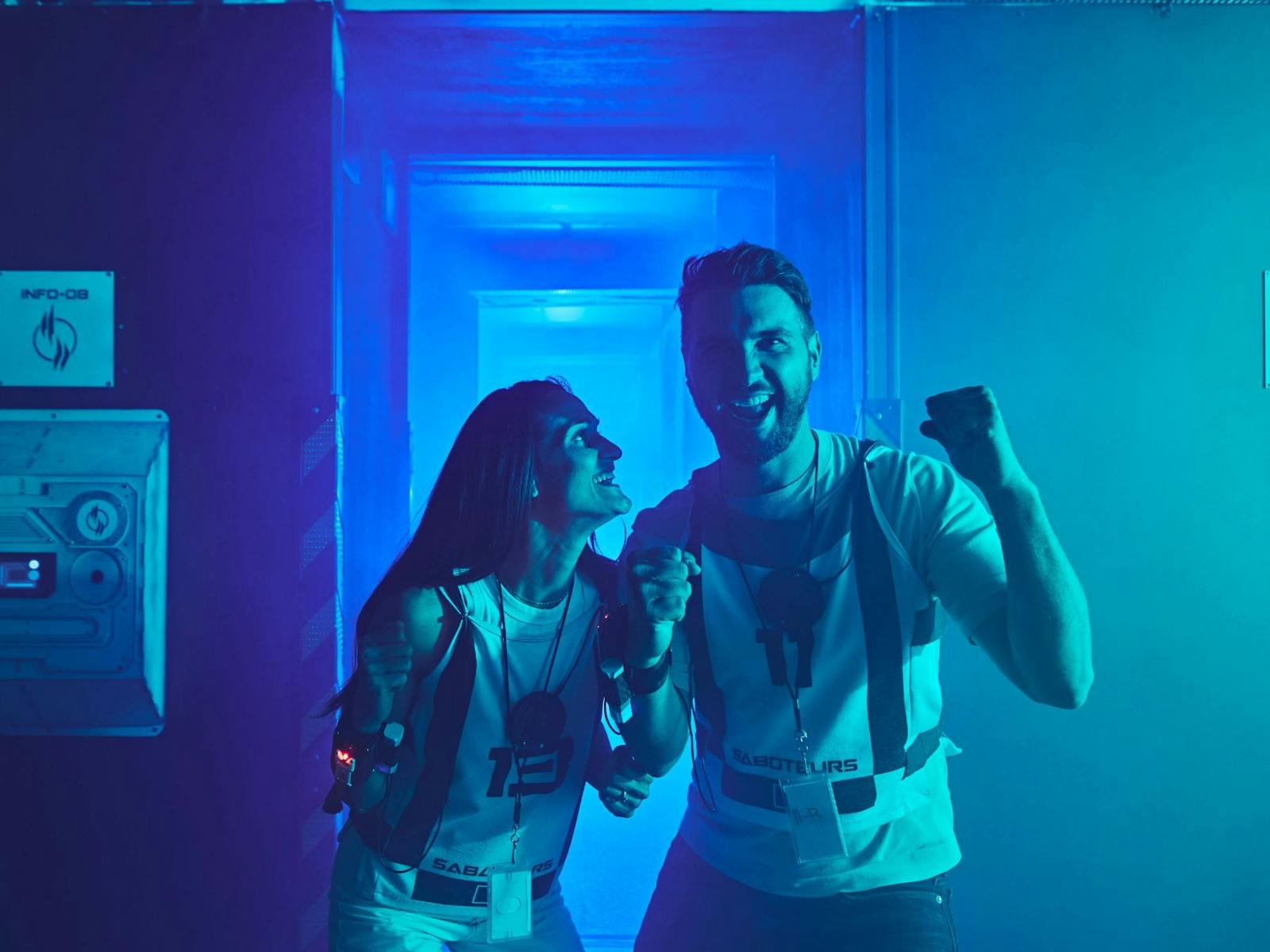 A man and woman in Saboteurs vests celebrate and laugh together in a blue-lit corridor. A man and woman in Saboteurs vests celebrate and laugh together in a blue-lit corridor.