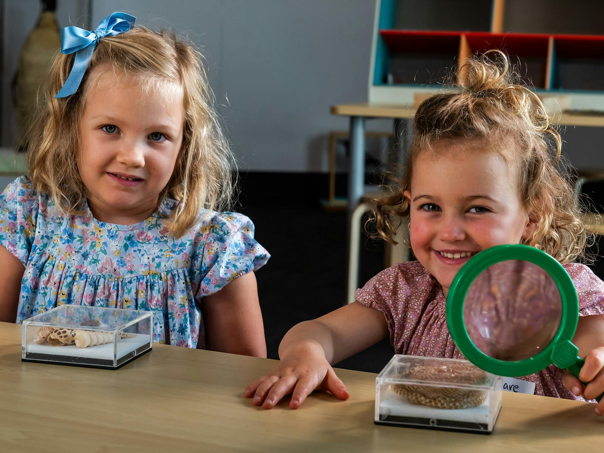 Two children smiling at camera. One holds a magnifiying glass and each have an acrylic seashell box