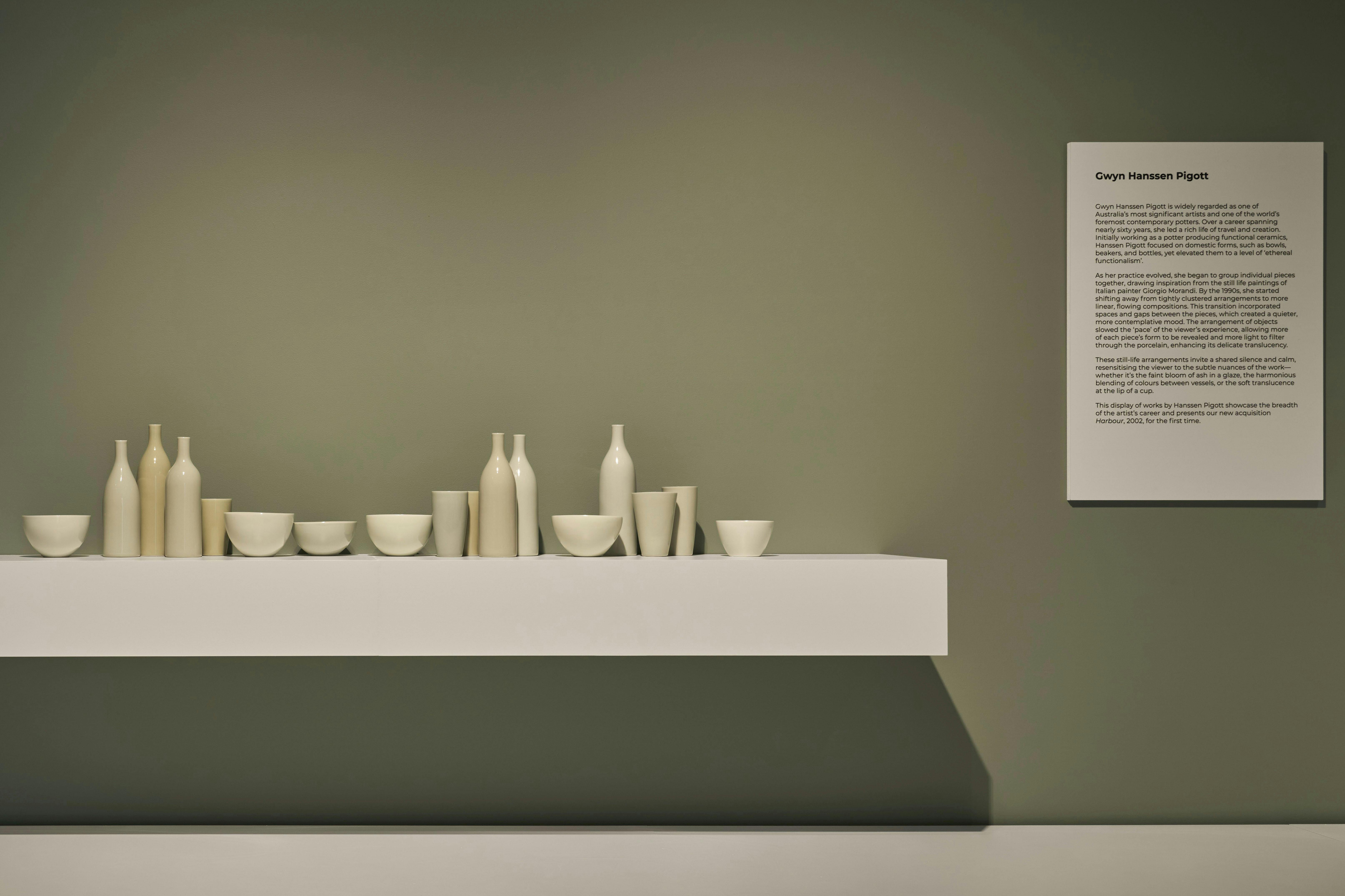 A row of pale porcelain vessels are displayed on a white shelf against a green wall in a gallery