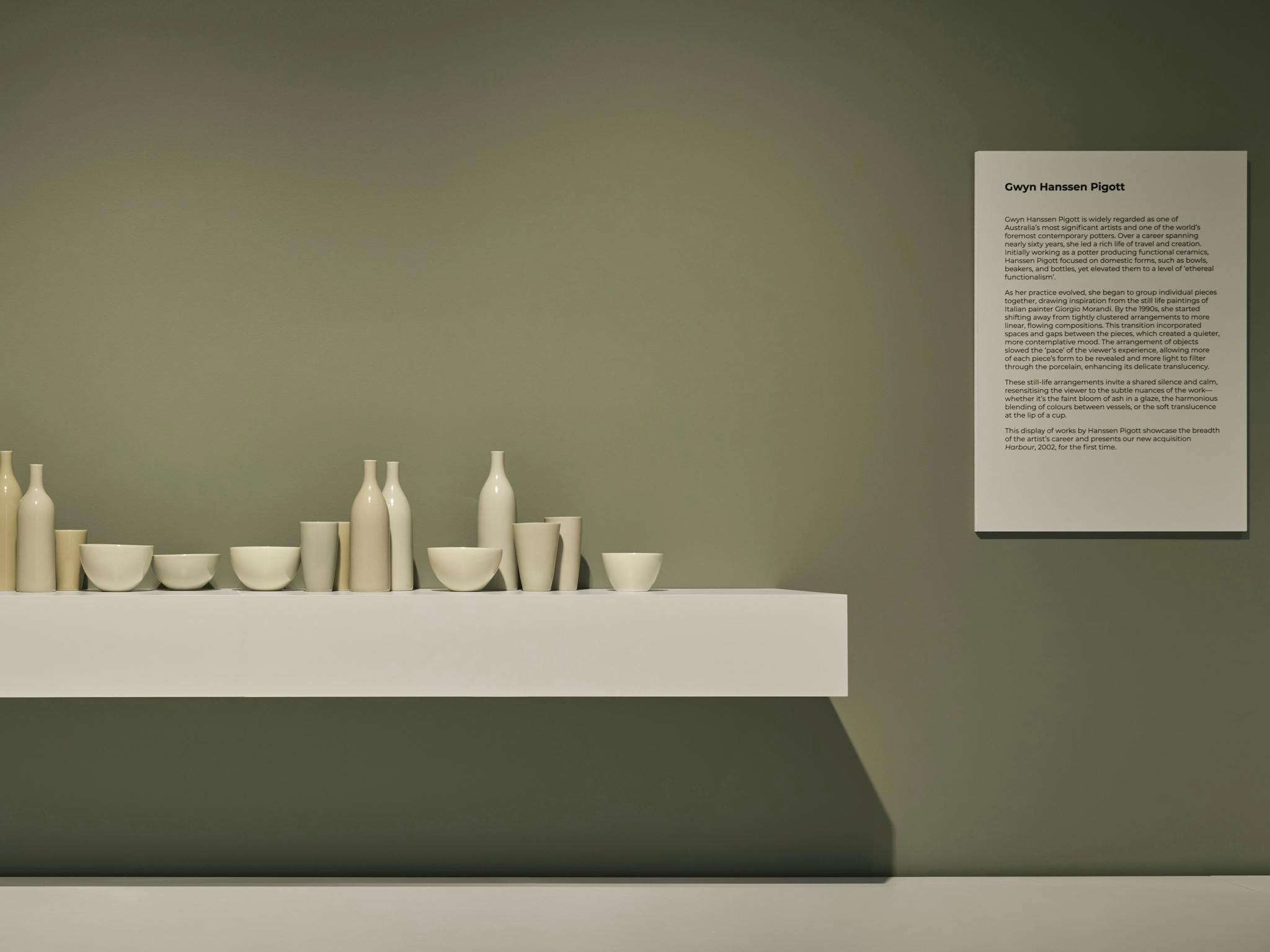 A row of pale porcelain vessels are displayed on a white shelf against a green wall in a gallery