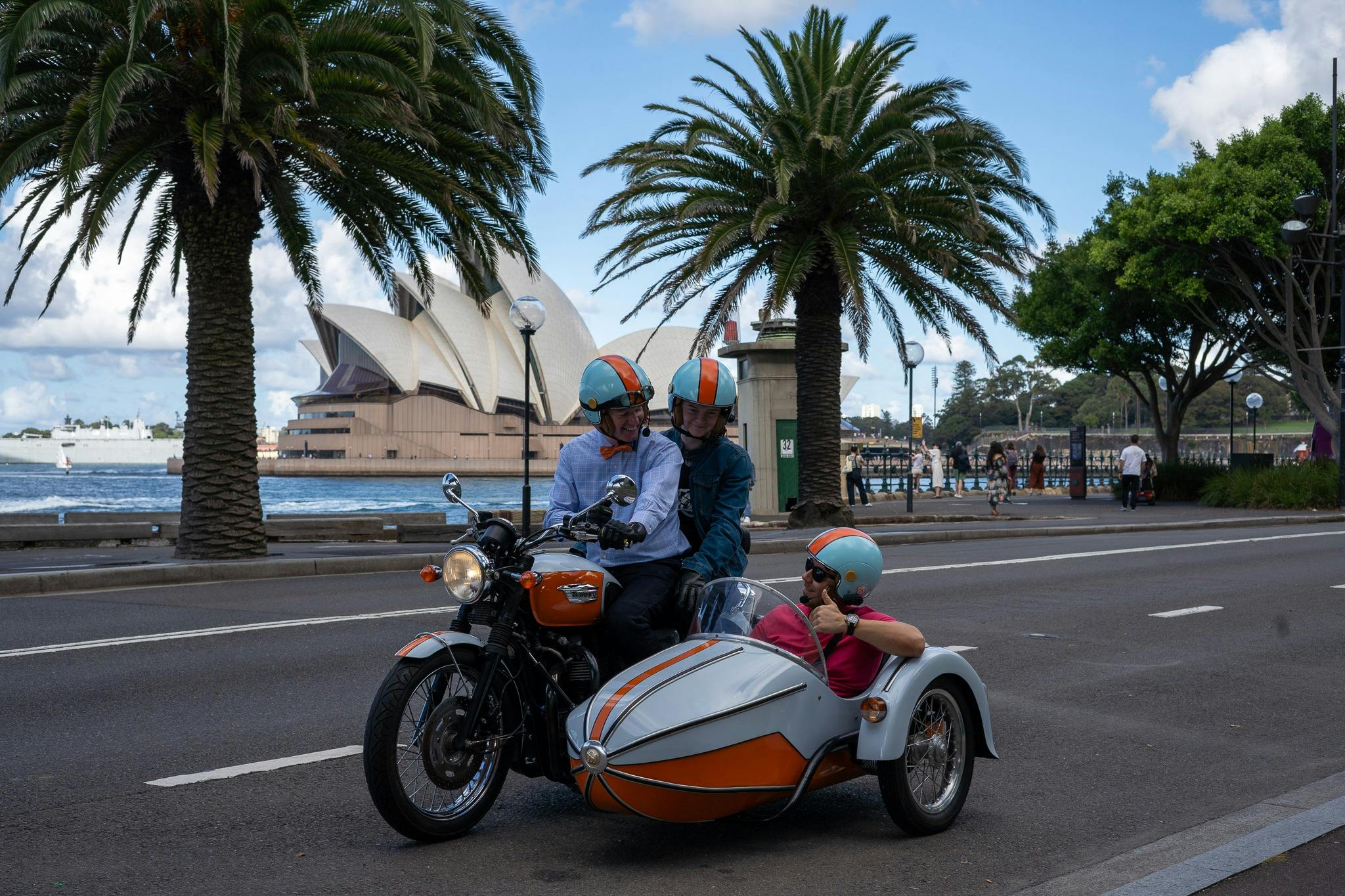 A motorcyle and sidecar across the water from the Sydney Opera House, with 3 smiling people onboard