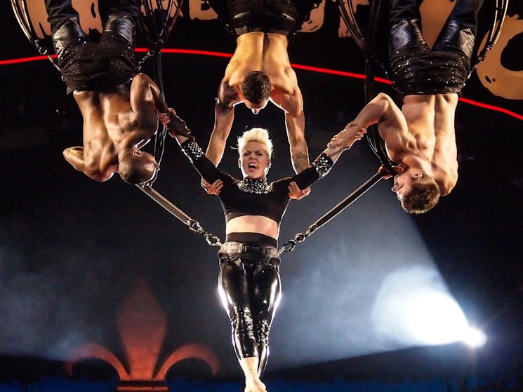 image shows the artist Pink hanging above the stage supported by 3 aerial performers holding her whi
