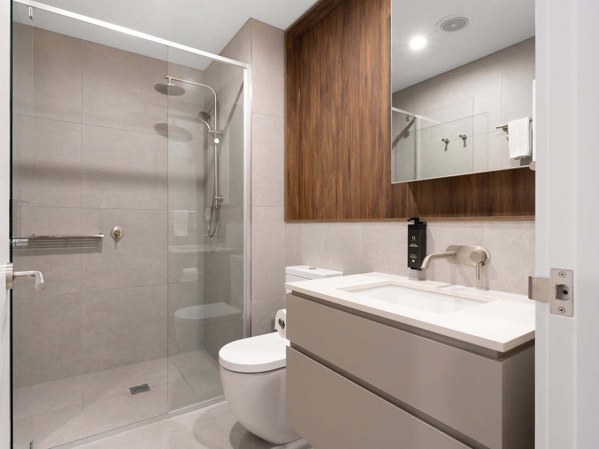 Modern bathroom with walk-in shower, toilet and sink. Grey and wood cabinetry with large mirror.