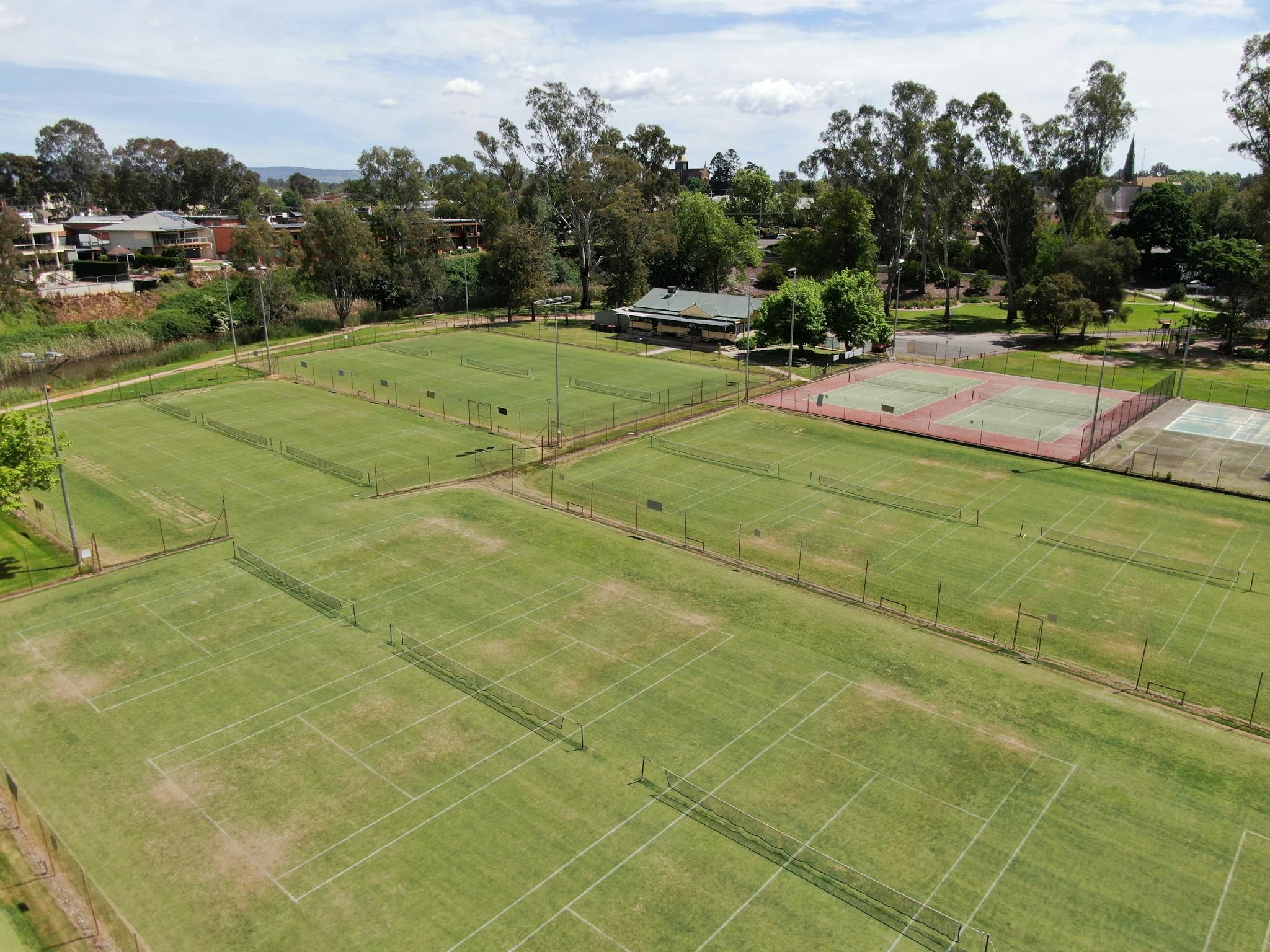 Aerial view of tennis courts, looking towards clubhouse, houses in distance, trees, sunny day.