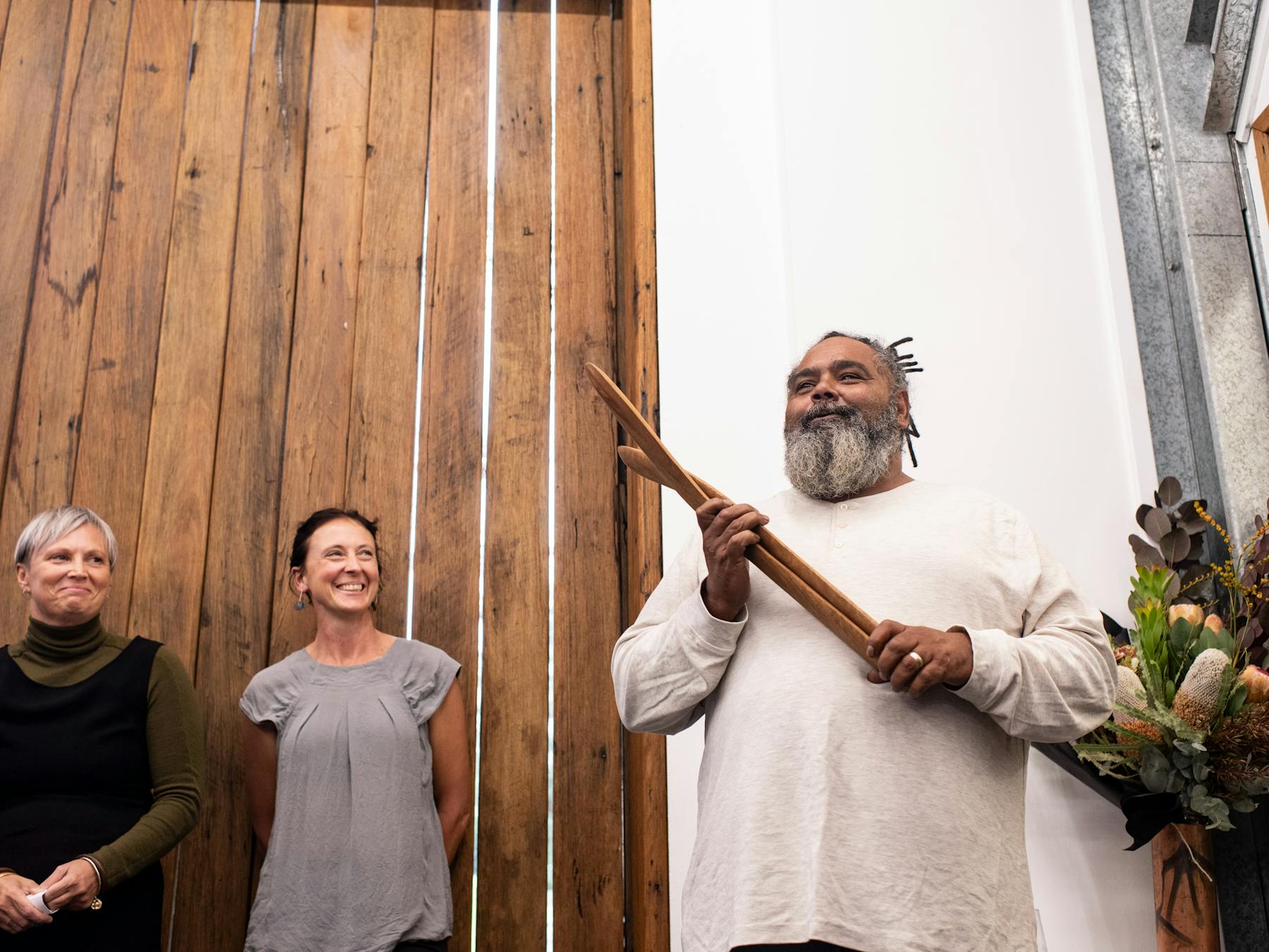 Aboriginal Artist holding wooden artefact next to a display of flowers with lady looking on smiling