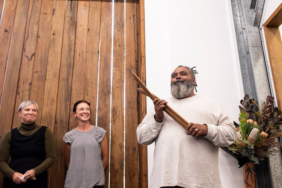 Aboriginal Artist holding wooden artefact next to a display of flowers with lady looking on smiling